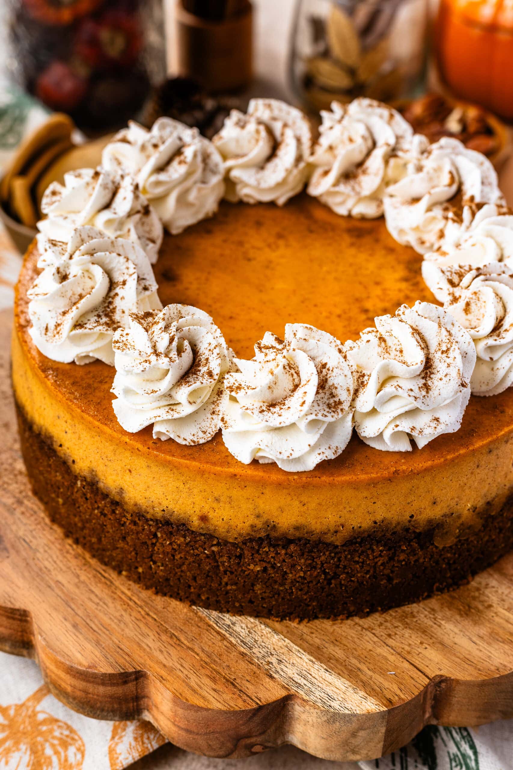 A decorated pumpkin cheesecake topped with whipped cream rosettes sprinkled with cinnamon, arranged in a circle around the edge, placed on a wooden board with cozy fall-themed decorations, including cookies, nuts, and a ceramic pumpkin in the background.