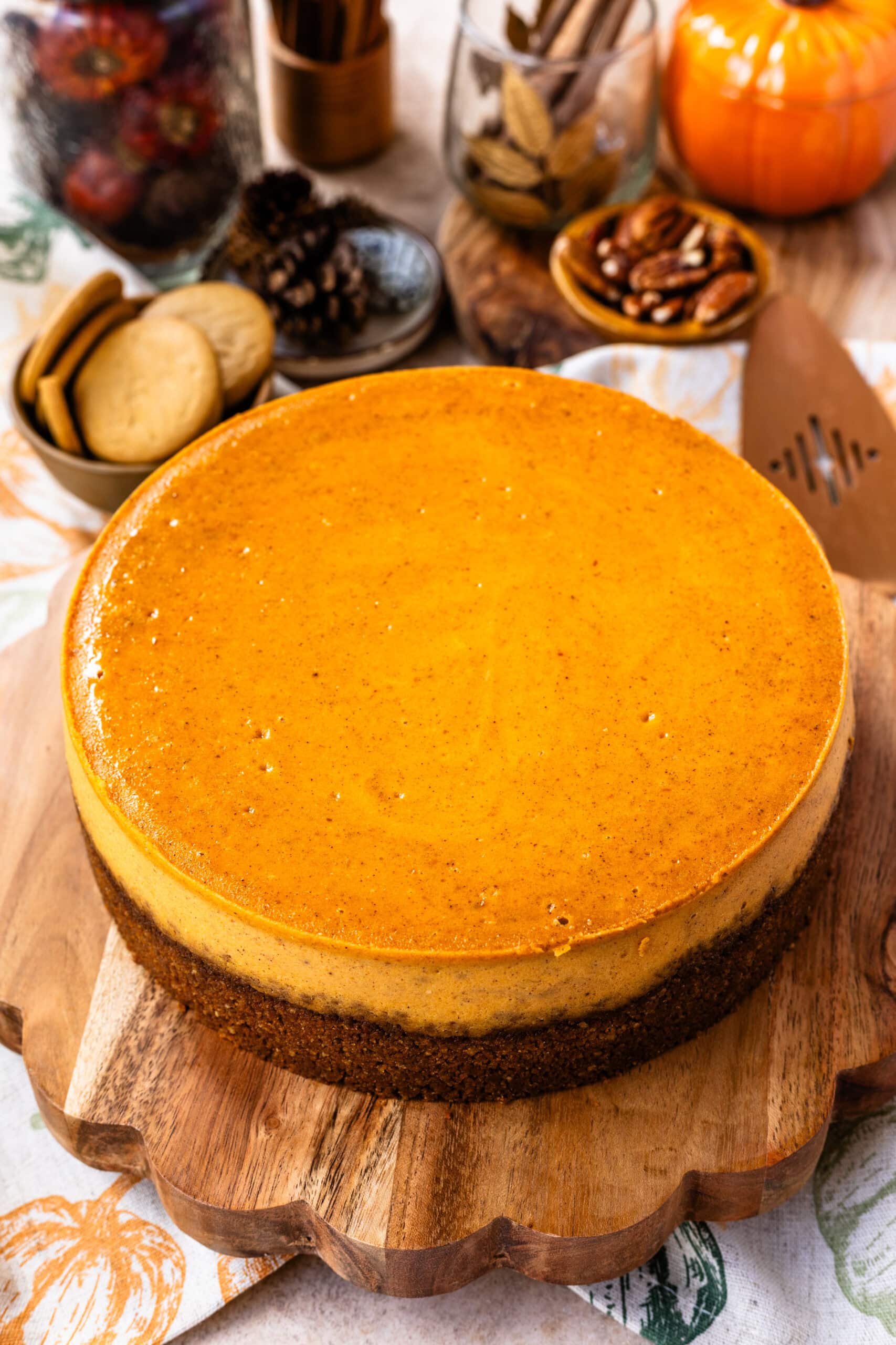 A close-up of a plain pumpkin cheesecake with a smooth orange surface and a graham cracker crust, sitting on a wooden board with fall decorations, including cookies, nuts, and cinnamon sticks, in the background.