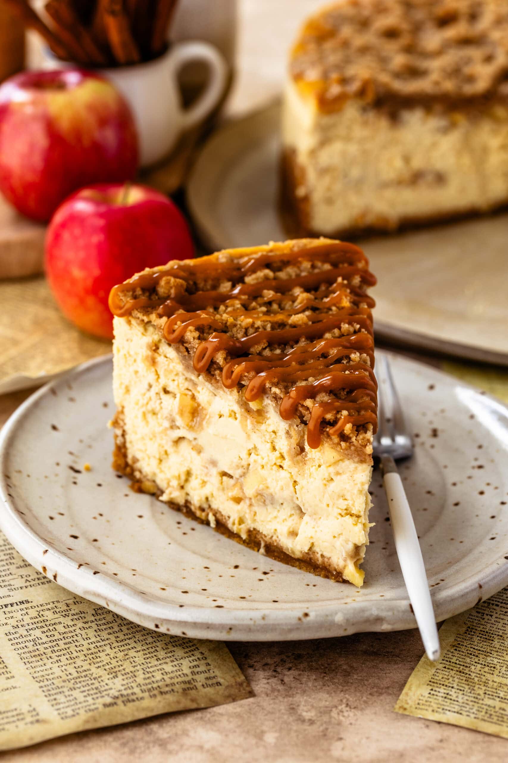 Close-up image of a slice of apple crumble cheesecake on a speckled ceramic plate, topped with a drizzle of caramel and a crumbly streusel. The cheesecake slice shows visible chunks of baked apples within the creamy filling. In the background, whole apples and the rest of the cheesecake are slightly blurred, adding a cozy and autumnal feel to the setting.