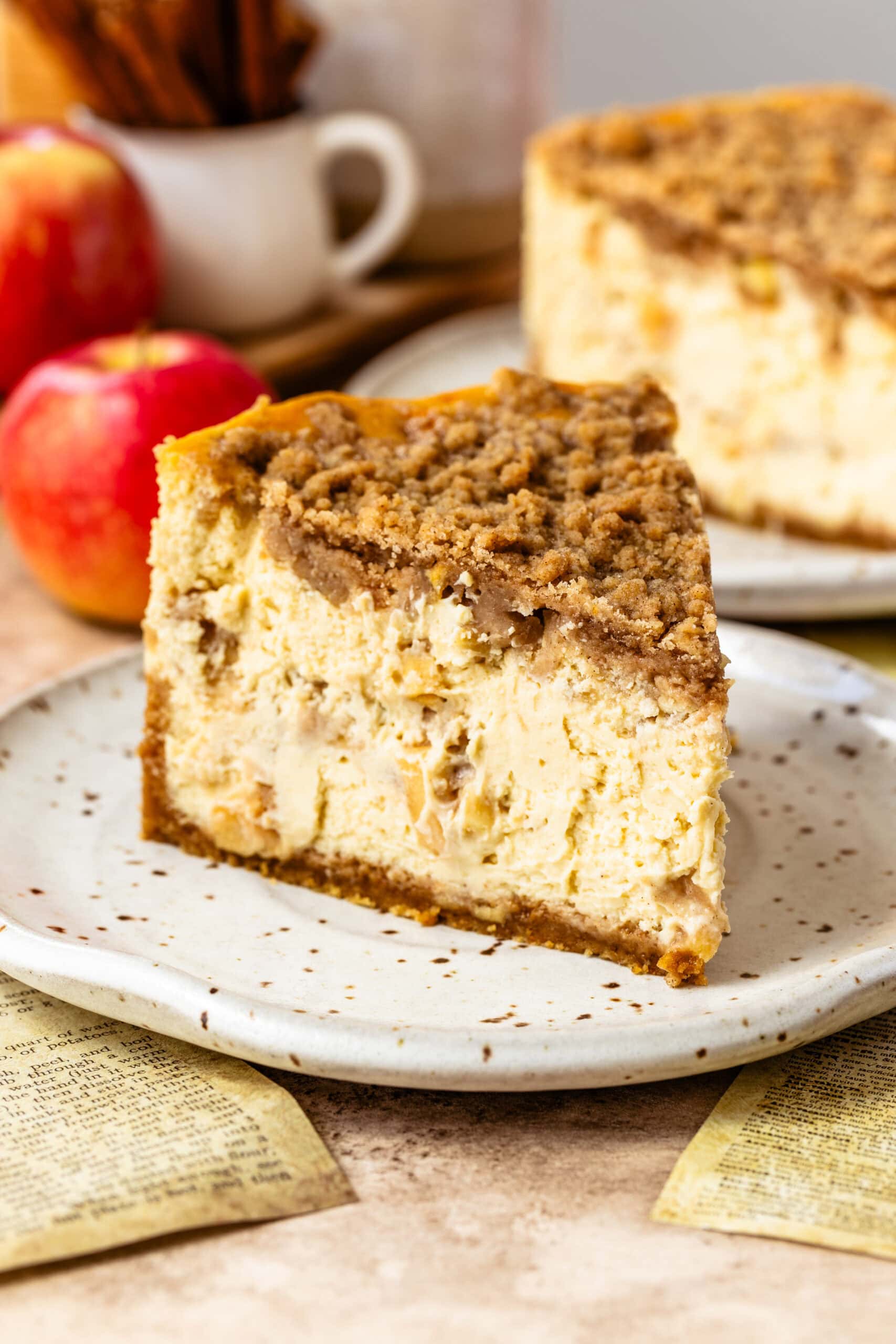 Close-up image of a slice of apple crumble cheesecake on a speckled ceramic plate. The cheesecake features visible apple chunks and a thick layer of crumble topping. The creamy texture of the cheesecake contrasts with the crunchy crumble. In the background, out of focus, are whole apples and the rest of the cheesecake on a plate, enhancing the rustic and warm autumnal atmosphere.