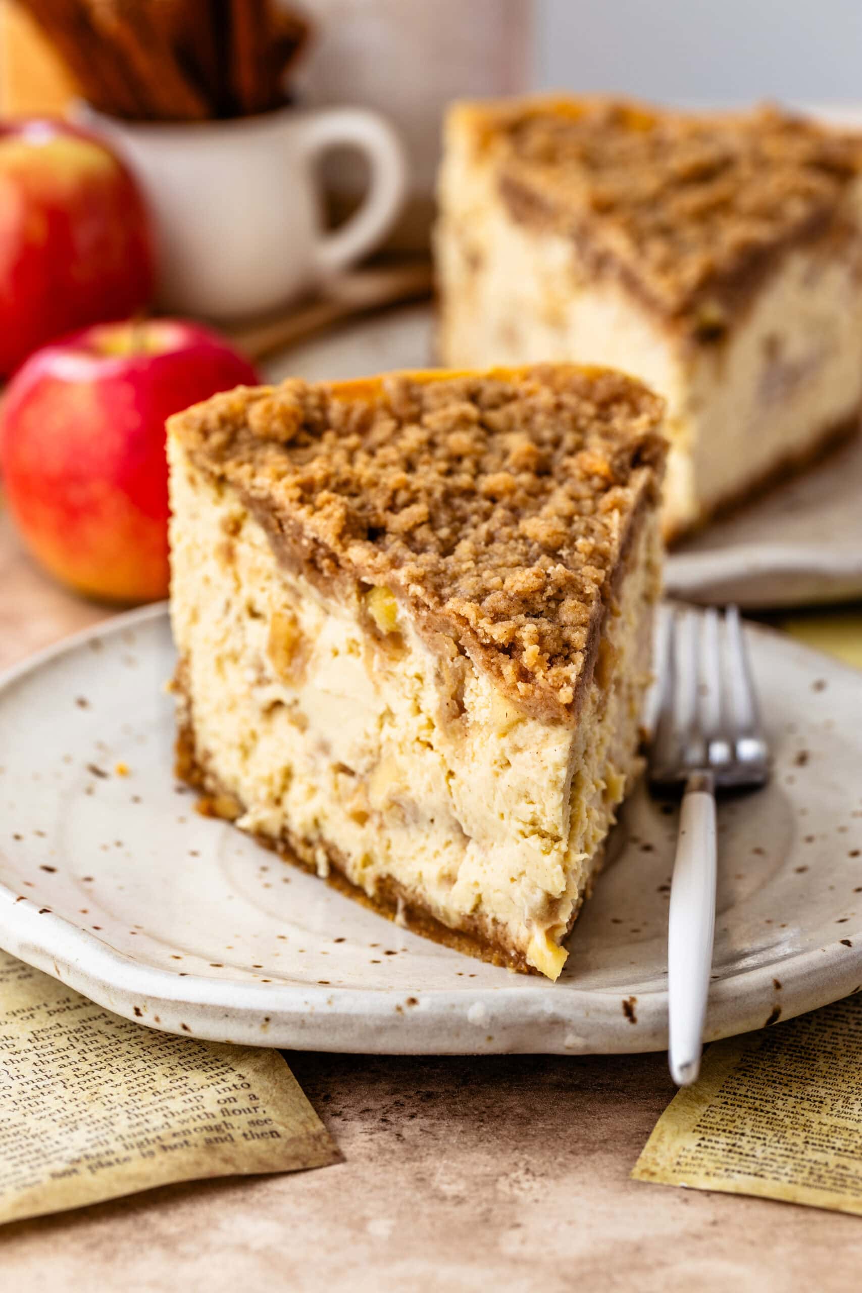 Close-up image of a slice of apple crumble cheesecake on a speckled ceramic plate. The cheesecake features visible apple chunks and a thick layer of crumble topping. The creamy texture of the cheesecake contrasts with the crunchy crumble. In the background, out of focus, are whole apples and the rest of the cheesecake on a plate, enhancing the rustic and warm autumnal atmosphere.
