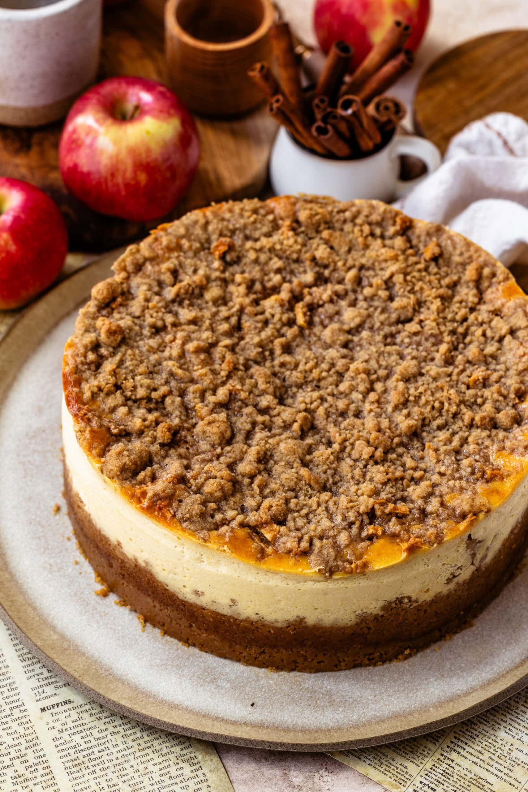 Close-up image of a freshly baked apple crumble cheesecake with a golden brown cinnamon crumble topping. The cheesecake features a creamy filling and a graham cracker crust, with apples visible in the filling beneath the crumble. The background includes apples and rustic kitchen items, complementing the autumnal theme of the dessert.