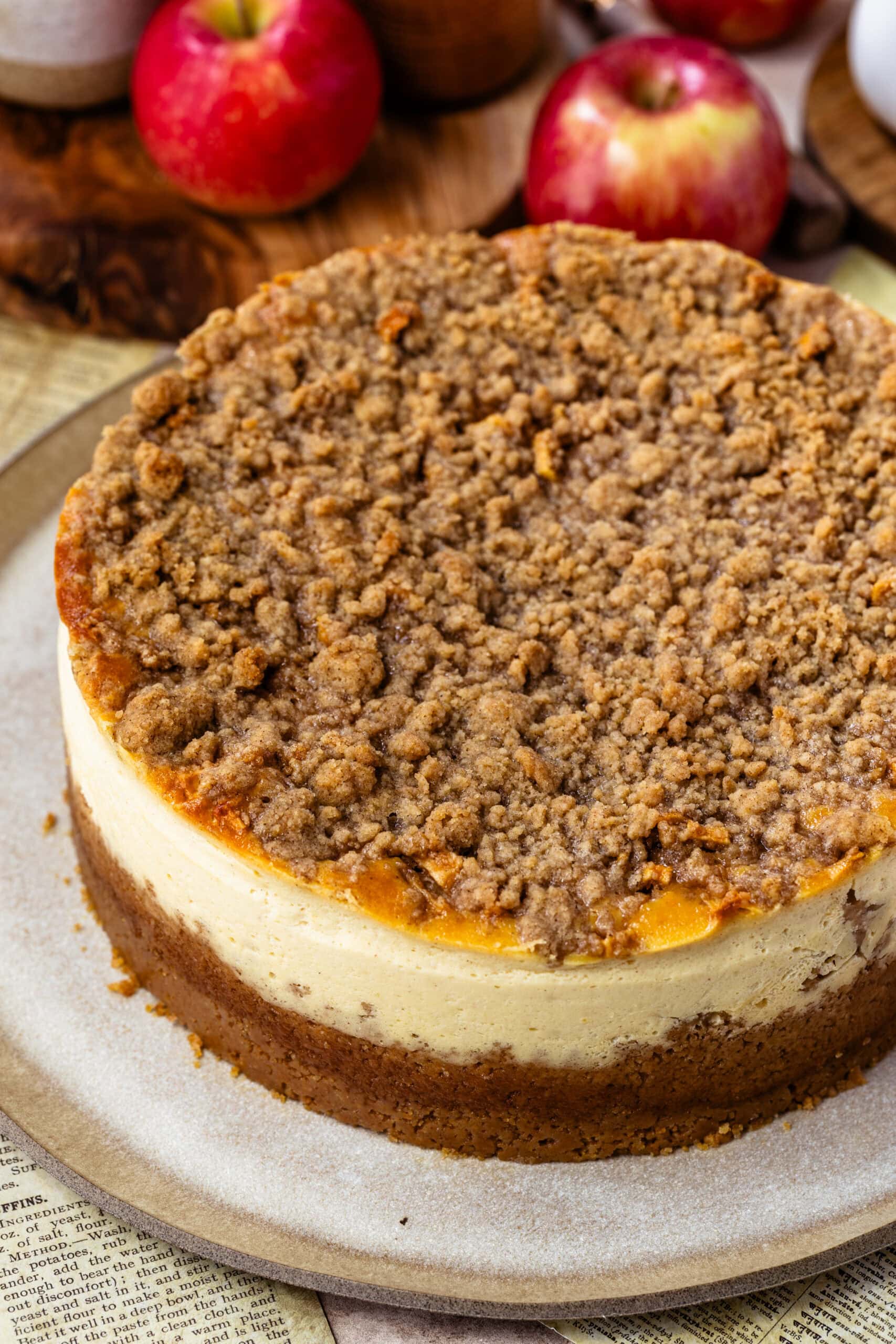 Close-up image of a freshly baked apple crumble cheesecake with a golden brown cinnamon crumble topping. The cheesecake features a creamy filling and a graham cracker crust, with apples visible in the filling beneath the crumble. The background includes apples and rustic kitchen items, complementing the autumnal theme of the dessert.