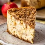 Close-up image of a slice of apple crumble cheesecake on a speckled ceramic plate. The cheesecake features visible apple chunks and a thick layer of crumble topping. The creamy texture of the cheesecake contrasts with the crunchy crumble. In the background, out of focus, are whole apples and the rest of the cheesecake on a plate, enhancing the rustic and warm autumnal atmosphere.