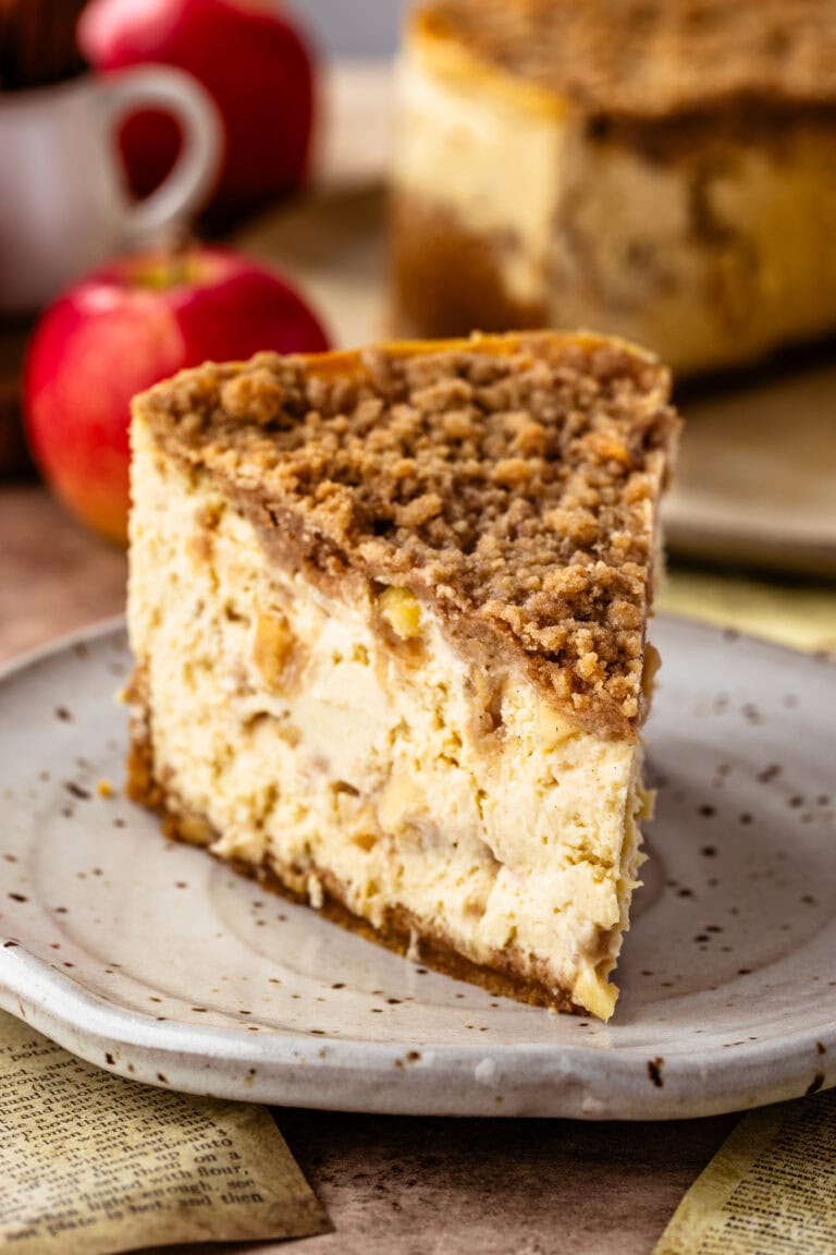 Close-up image of a slice of apple crumble cheesecake on a speckled ceramic plate. The cheesecake features visible apple chunks and a thick layer of crumble topping. The creamy texture of the cheesecake contrasts with the crunchy crumble. In the background, out of focus, are whole apples and the rest of the cheesecake on a plate, enhancing the rustic and warm autumnal atmosphere.