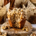 A close-up of a banana nut muffin with a golden-brown exterior, topped with a crumbly walnut streusel. The muffin is placed on a wooden board with scattered walnut pieces around it, and there is a visible bite taken from the muffin, showing its soft interior.