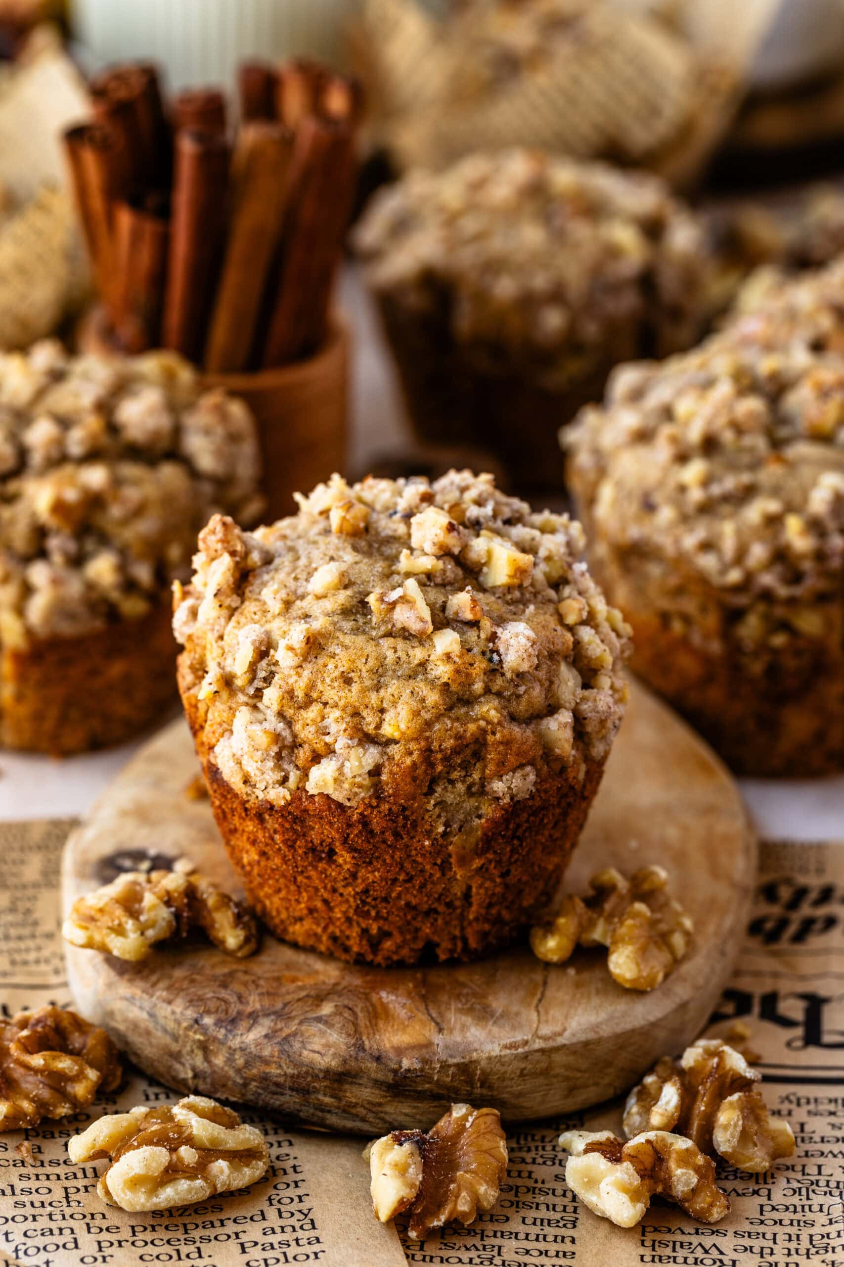 A banana nut muffin with a crispy walnut streusel topping, standing upright on a wooden board. The background features more muffins, cinnamon sticks, and scattered walnuts, creating a cozy, rustic atmosphere.