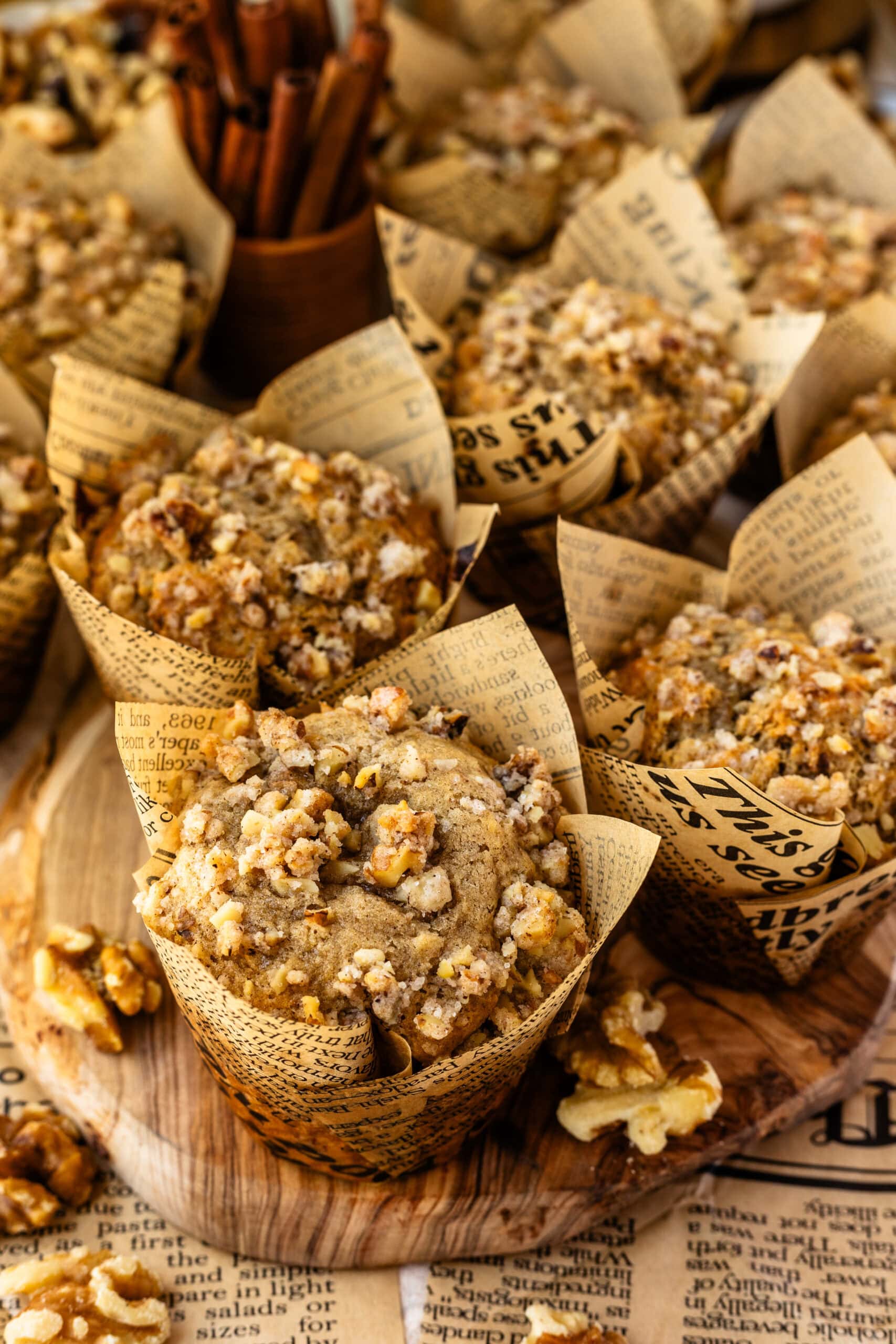 Multiple banana nut muffins wrapped in newspaper-style liners, arranged on a wooden board. Each muffin is topped with a generous portion of walnut streusel, giving them a crunchy appearance. In the background, cinnamon sticks and additional muffins are visible.