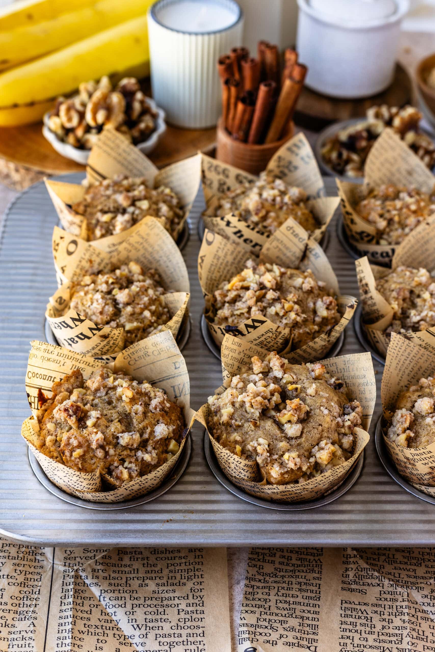 A muffin tray filled with freshly baked banana nut muffins, each wrapped in newspaper-patterned liners and topped with a crunchy walnut streusel. In the background, bananas, cinnamon sticks, and a small bowl of walnuts are arranged on a rustic table.