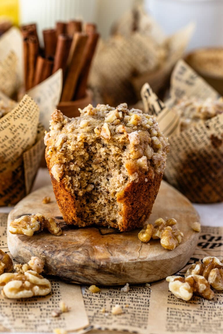 A close-up of a banana nut muffin with a golden-brown exterior, topped with a crumbly walnut streusel. The muffin is placed on a wooden board with scattered walnut pieces around it, and there is a visible bite taken from the muffin, showing its soft interior.