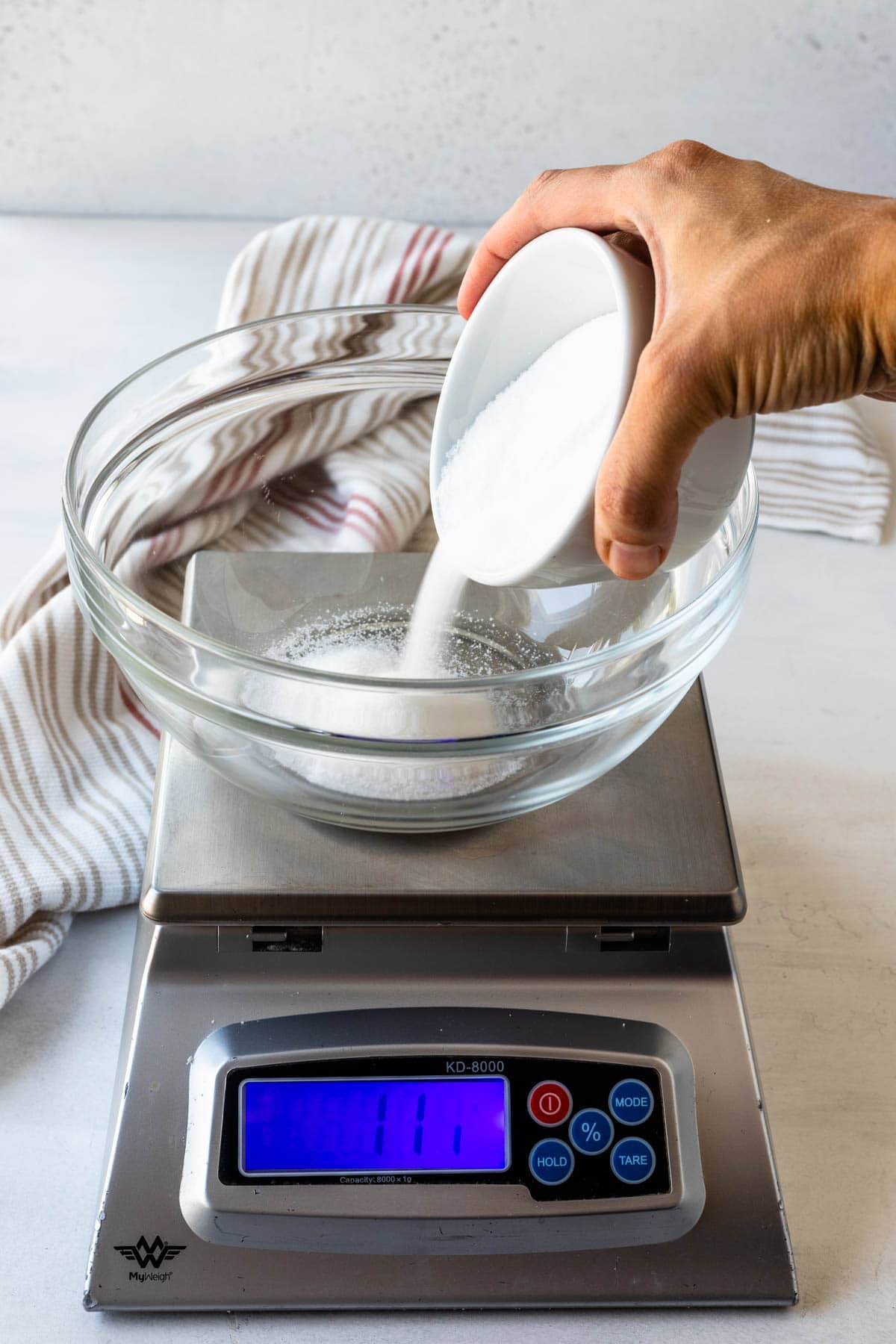 Hand pouring granulated sugar from a small bowl into a larger glass bowl placed on a digital kitchen scale, with the display showing 29 grams.