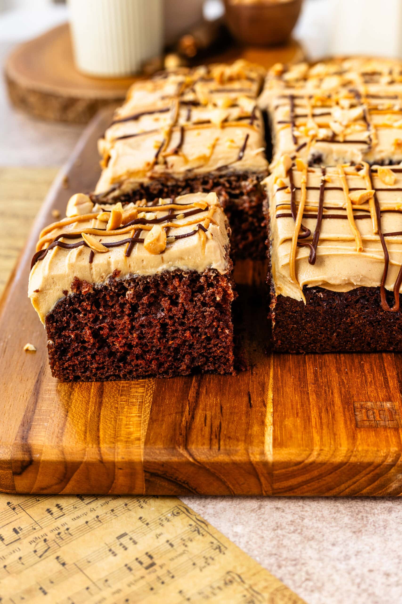 Chocolate Cake with Peanut Butter Frosting Cut into Squares: A close-up image of a chocolate cake with peanut butter frosting, cut into squares and served on a wooden cutting board. The frosting is topped with a drizzle of chocolate and peanut butter, and sprinkled with chopped peanuts. The focus is on the front slice, showing the rich, moist texture of the cake and the creamy frosting on top.