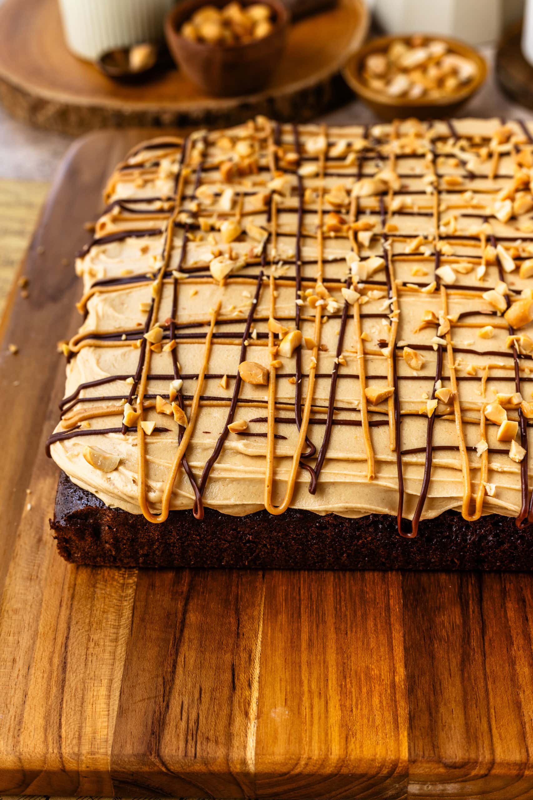 Whole Chocolate Cake with Peanut Butter Frosting: A whole chocolate cake topped with a generous layer of peanut butter frosting, drizzled with chocolate and peanut butter, and sprinkled with chopped peanuts. The cake is presented on a wooden board, with a cozy kitchen scene in the background, including bowls of peanuts and chocolate pieces.