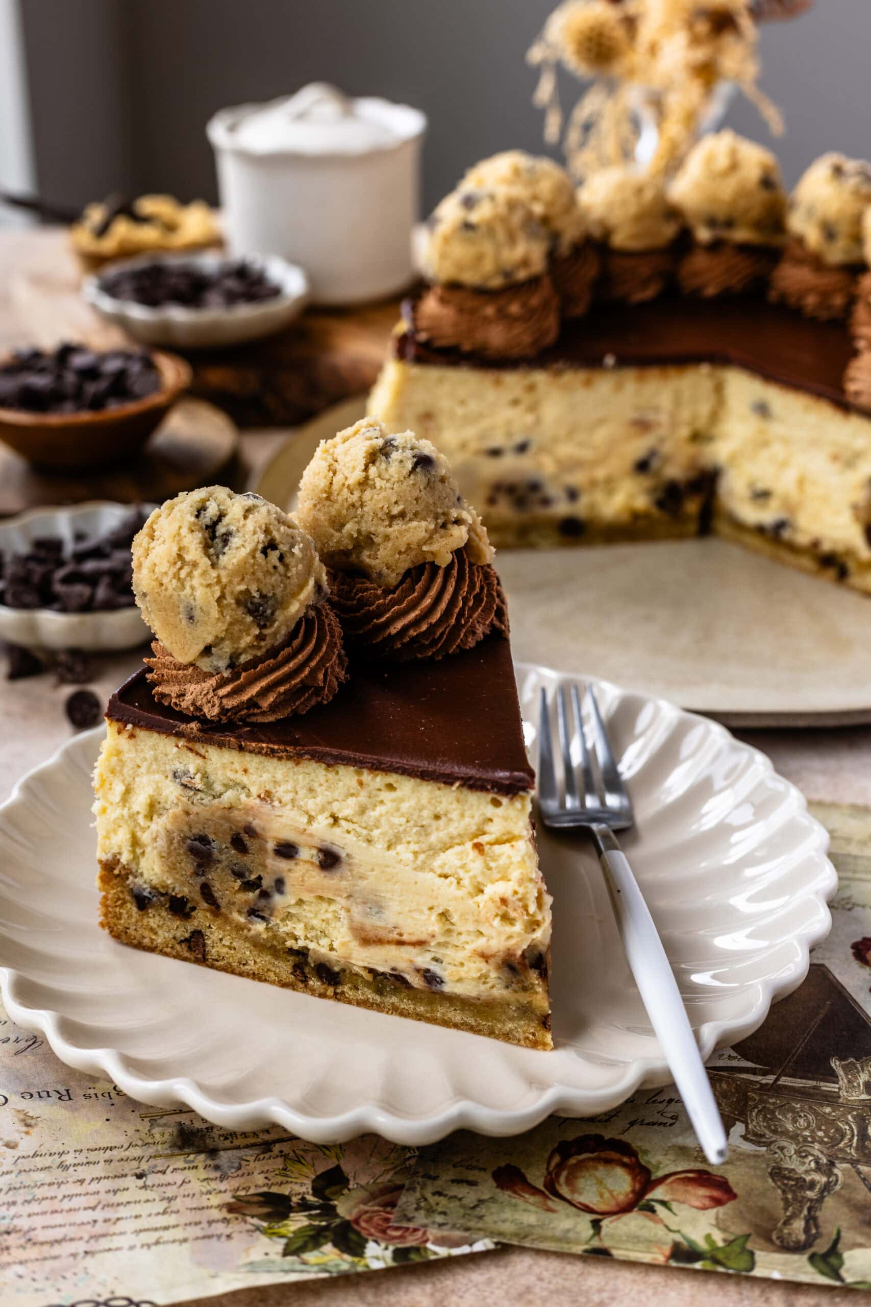 A slice of cookie dough cheesecake on a plate, showcasing layers of cheesecake, chocolate chip cookie dough, and a cookie crust, topped with chocolate ganache and garnished with chocolate frosting and cookie dough balls.