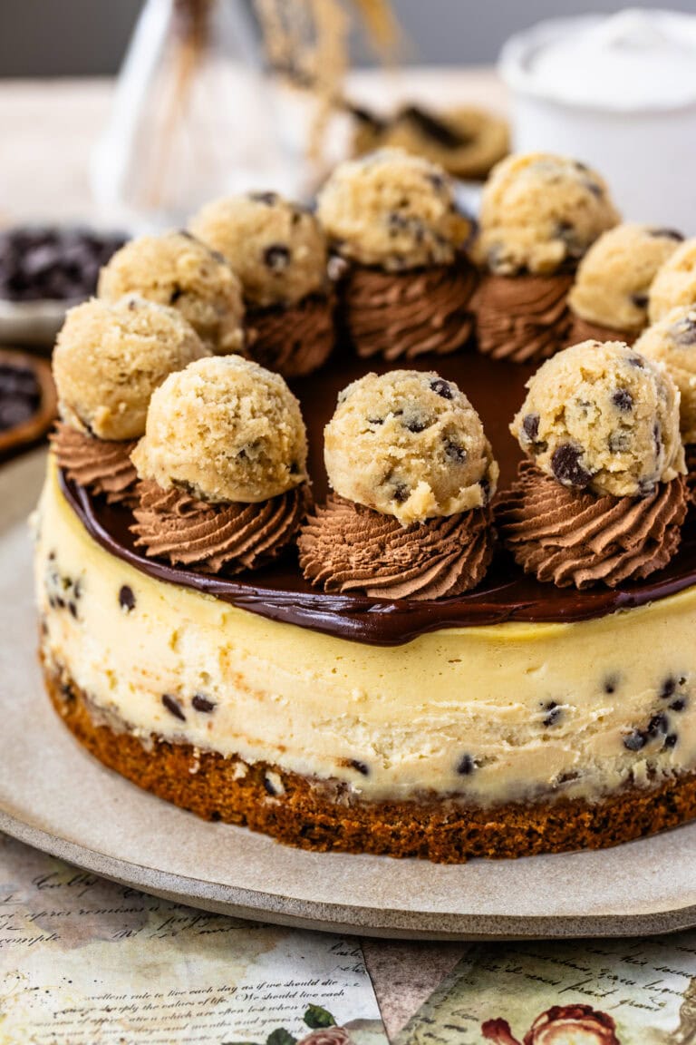 A close-up of a cookie dough cheesecake topped with rich chocolate ganache, swirls of chocolate frosting, and garnished with cookie dough balls, sitting on a cookie crust and studded with chocolate chips.