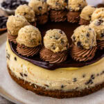 A close-up of a cookie dough cheesecake topped with rich chocolate ganache, swirls of chocolate frosting, and garnished with cookie dough balls, sitting on a cookie crust and studded with chocolate chips.