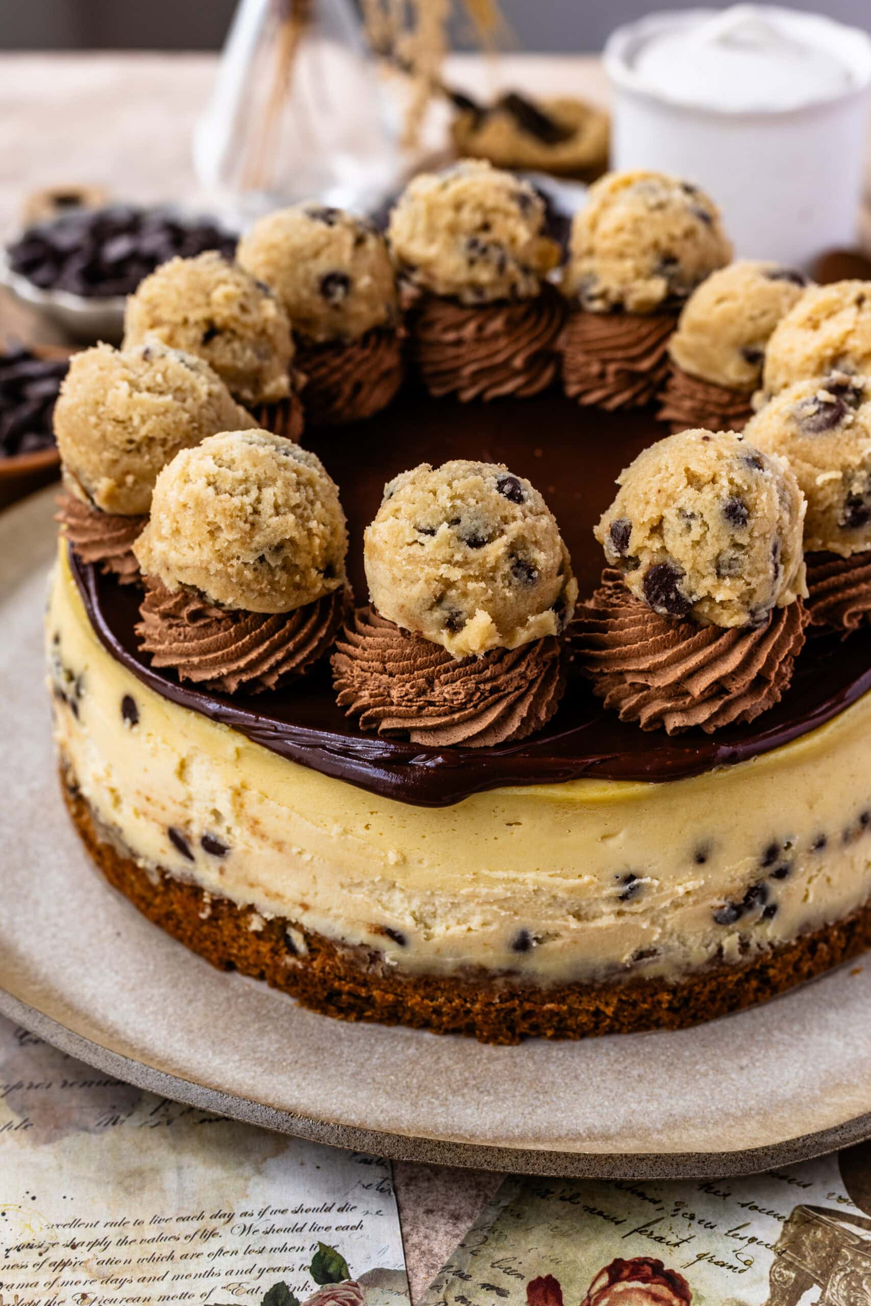 A close-up of a cookie dough cheesecake topped with rich chocolate ganache, swirls of chocolate frosting, and garnished with cookie dough balls, sitting on a cookie crust and studded with chocolate chips.