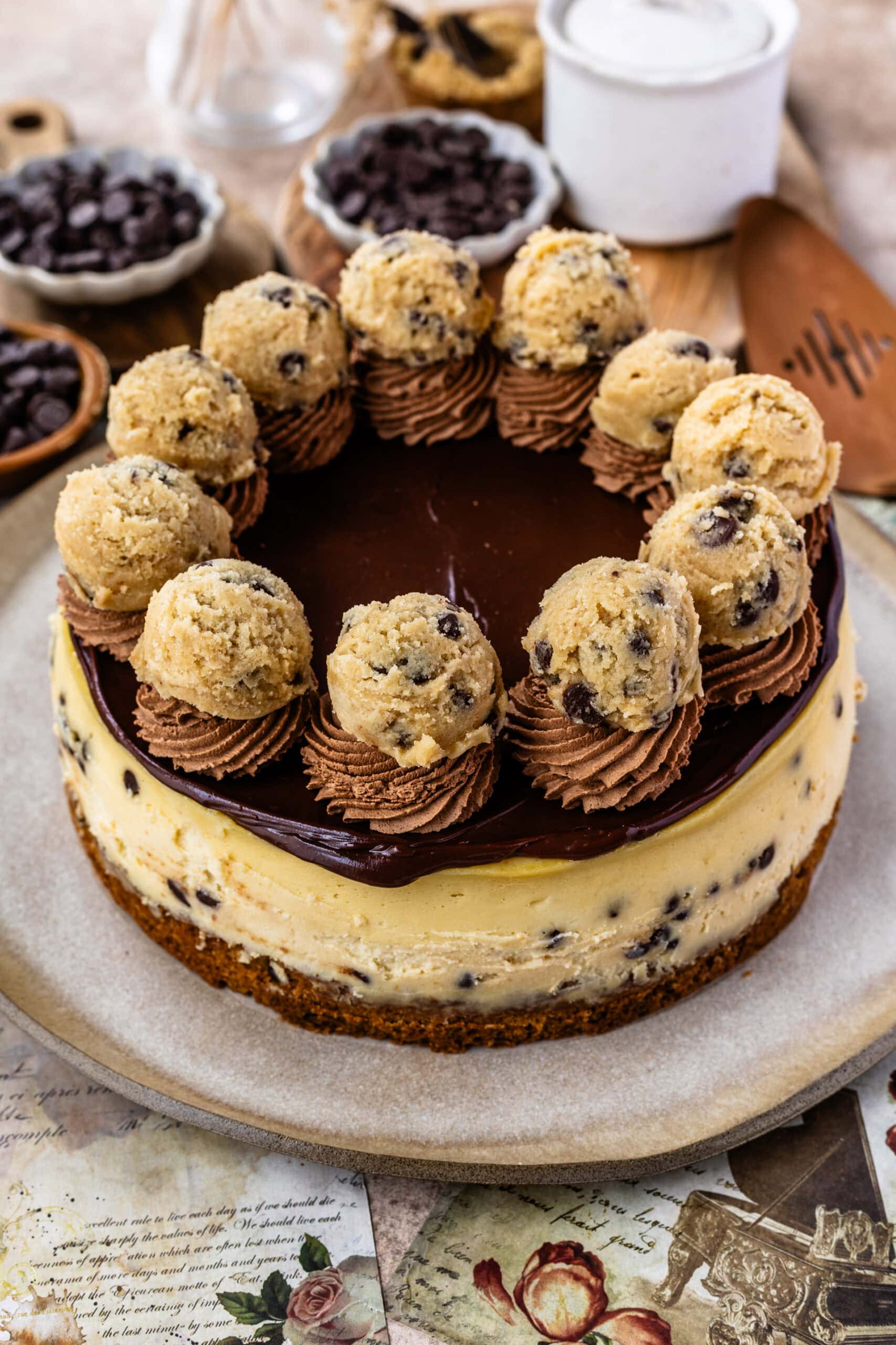 A close-up of a cookie dough cheesecake topped with rich chocolate ganache, swirls of chocolate frosting, and garnished with cookie dough balls, sitting on a cookie crust and studded with chocolate chips.