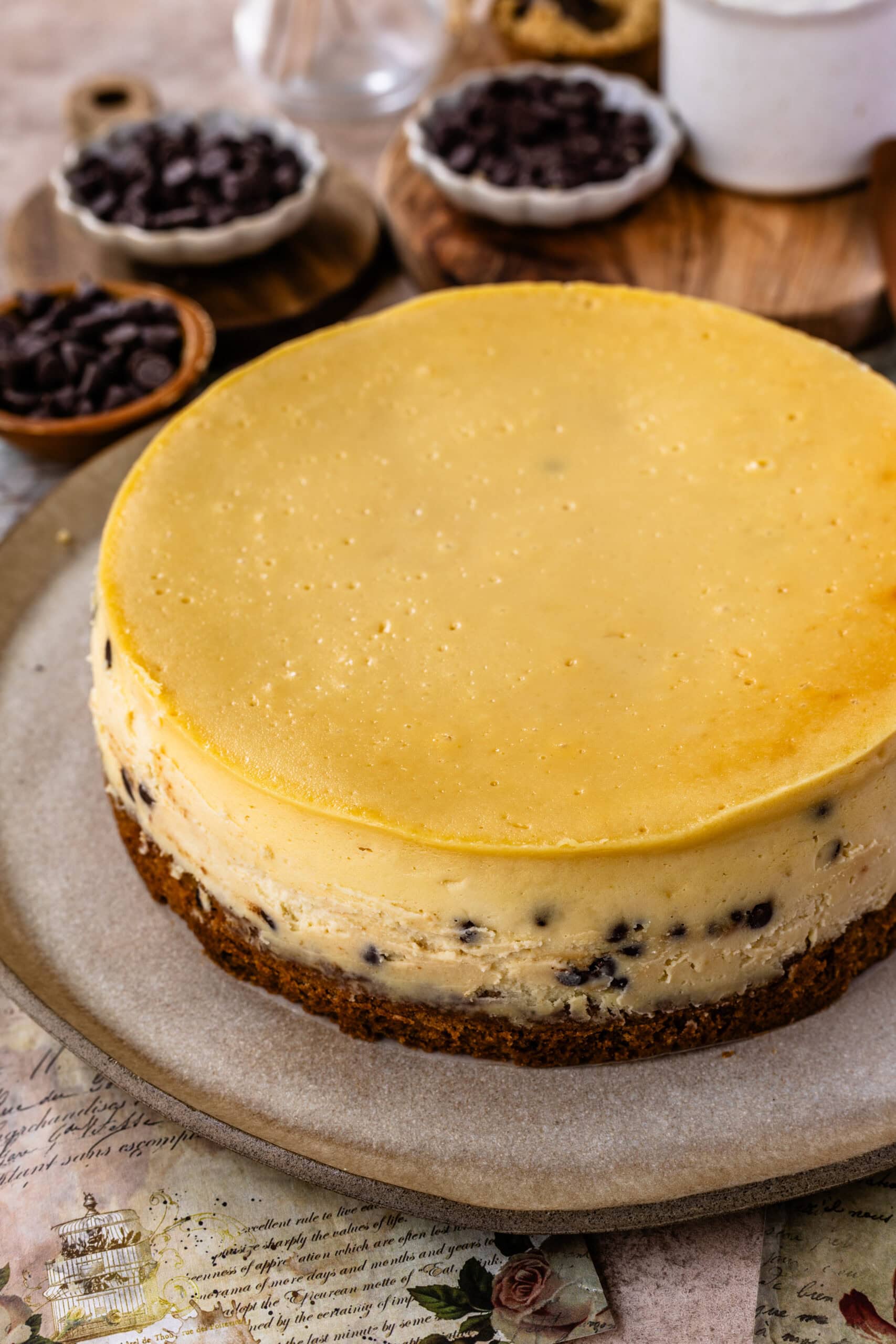 A plain baked cookie dough cheesecake with a smooth top and chocolate chip cookie crust, ready for decoration, displayed on a plate with bowls of chocolate chips visible in the background.