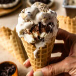 A hand holding a waffle cone filled with cookie dough ice cream, showcasing the swirls of fudge and cookie dough chunks.