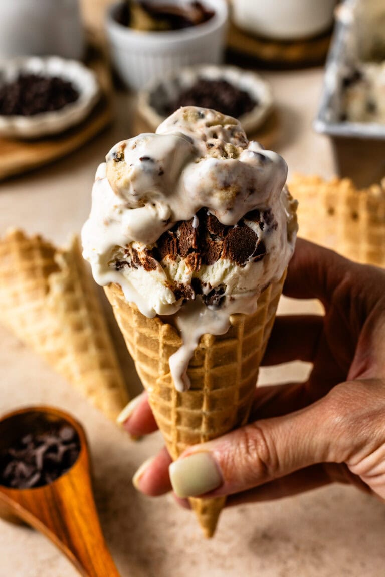 A hand holding a waffle cone filled with cookie dough ice cream, showcasing the swirls of fudge and cookie dough chunks.