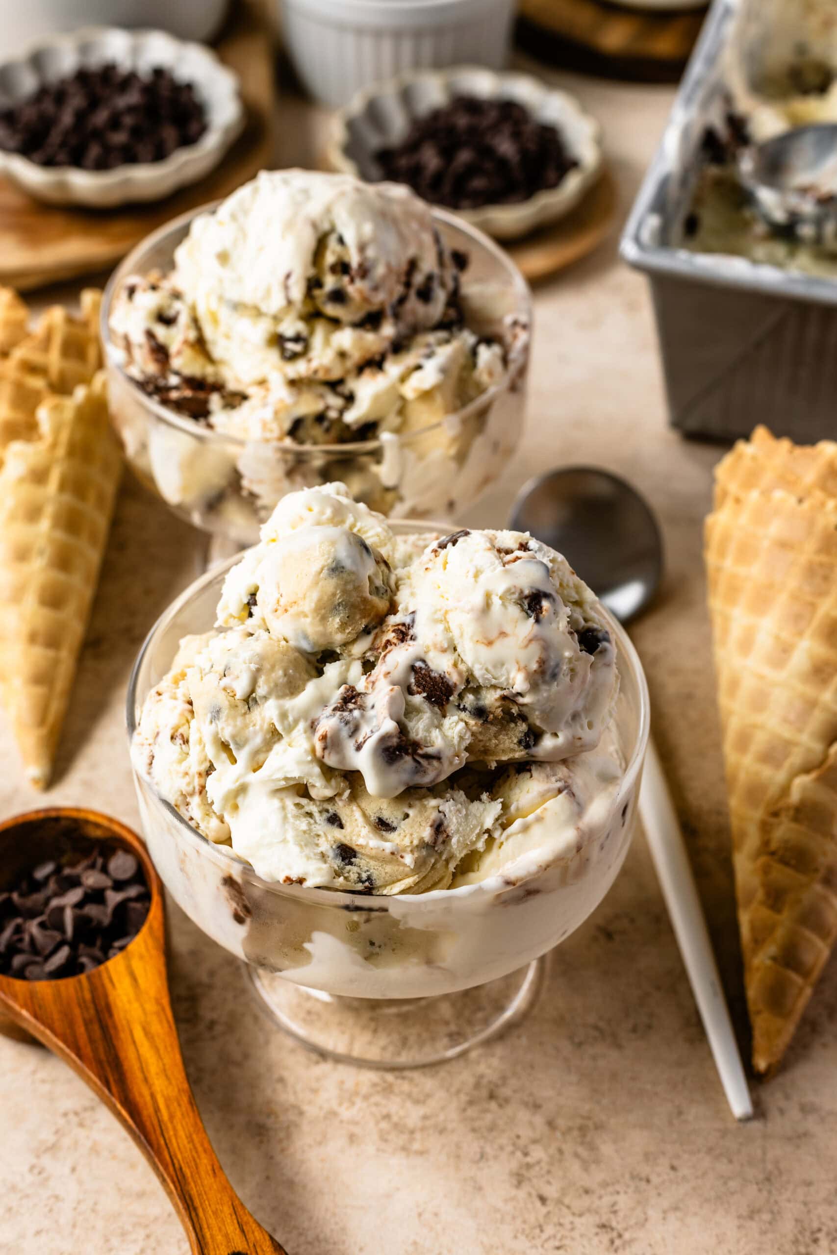 Two bowls filled with cookie dough ice cream, topped with chunks of cookie dough and chocolate chips, with waffle cones and chocolate chips in the background.