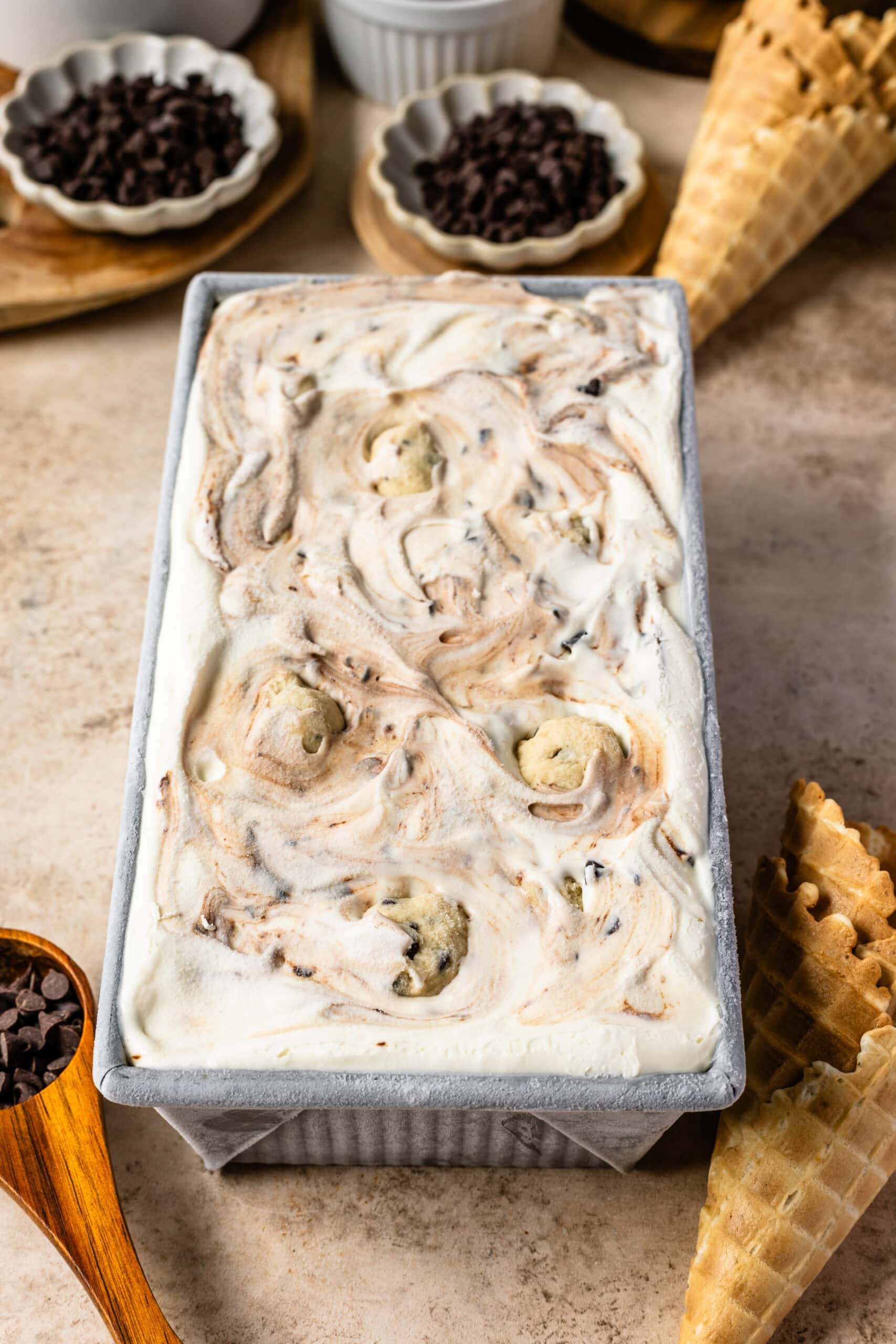 A loaf pan filled with homemade cookie dough ice cream, swirled with fudge and studded with cookie dough balls, placed on a countertop with waffle cones and chocolate chips in the background.