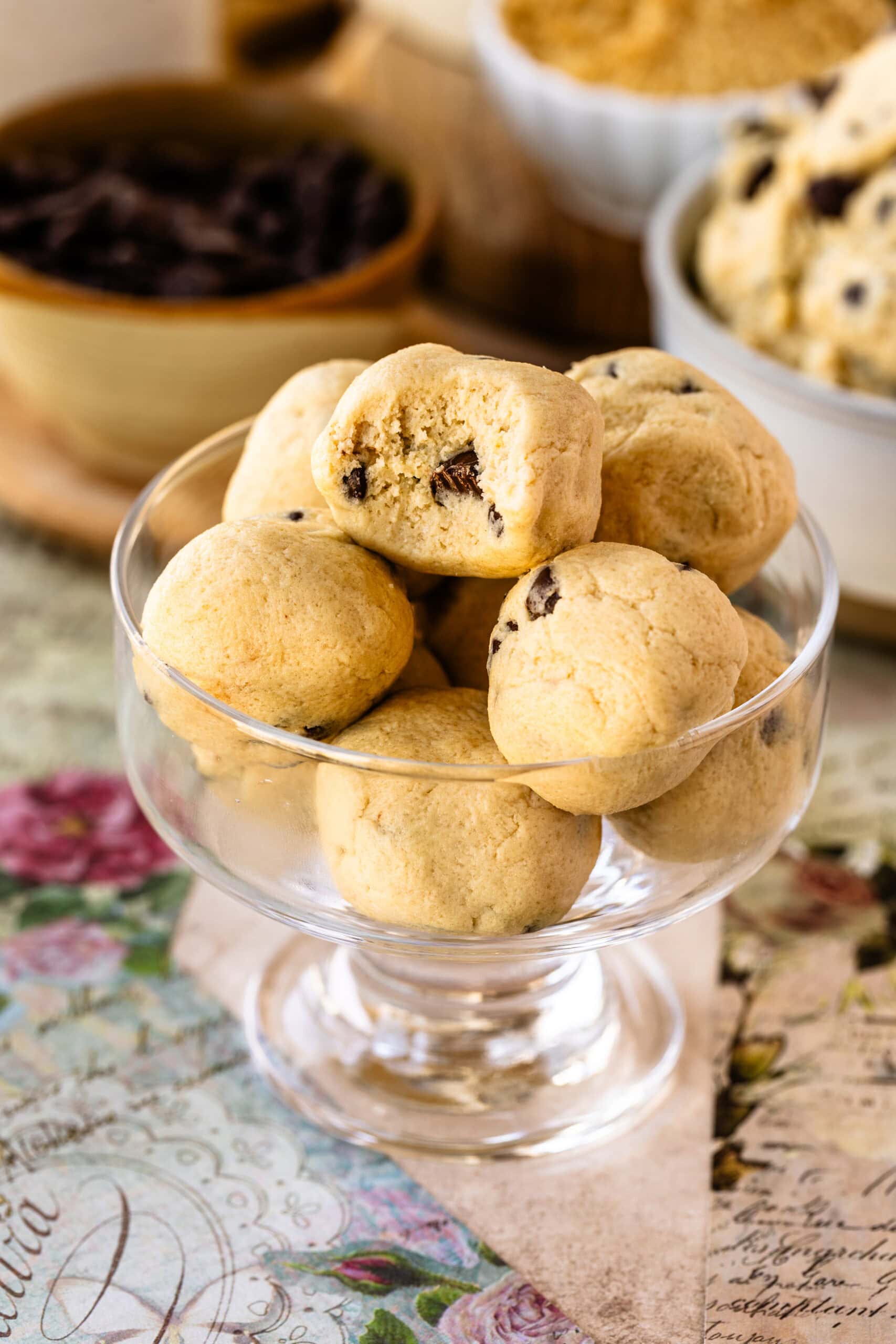 A glass dish filled with bite-sized balls of edible cookie dough, one with a bite taken out, revealing the soft texture and chocolate chips inside.