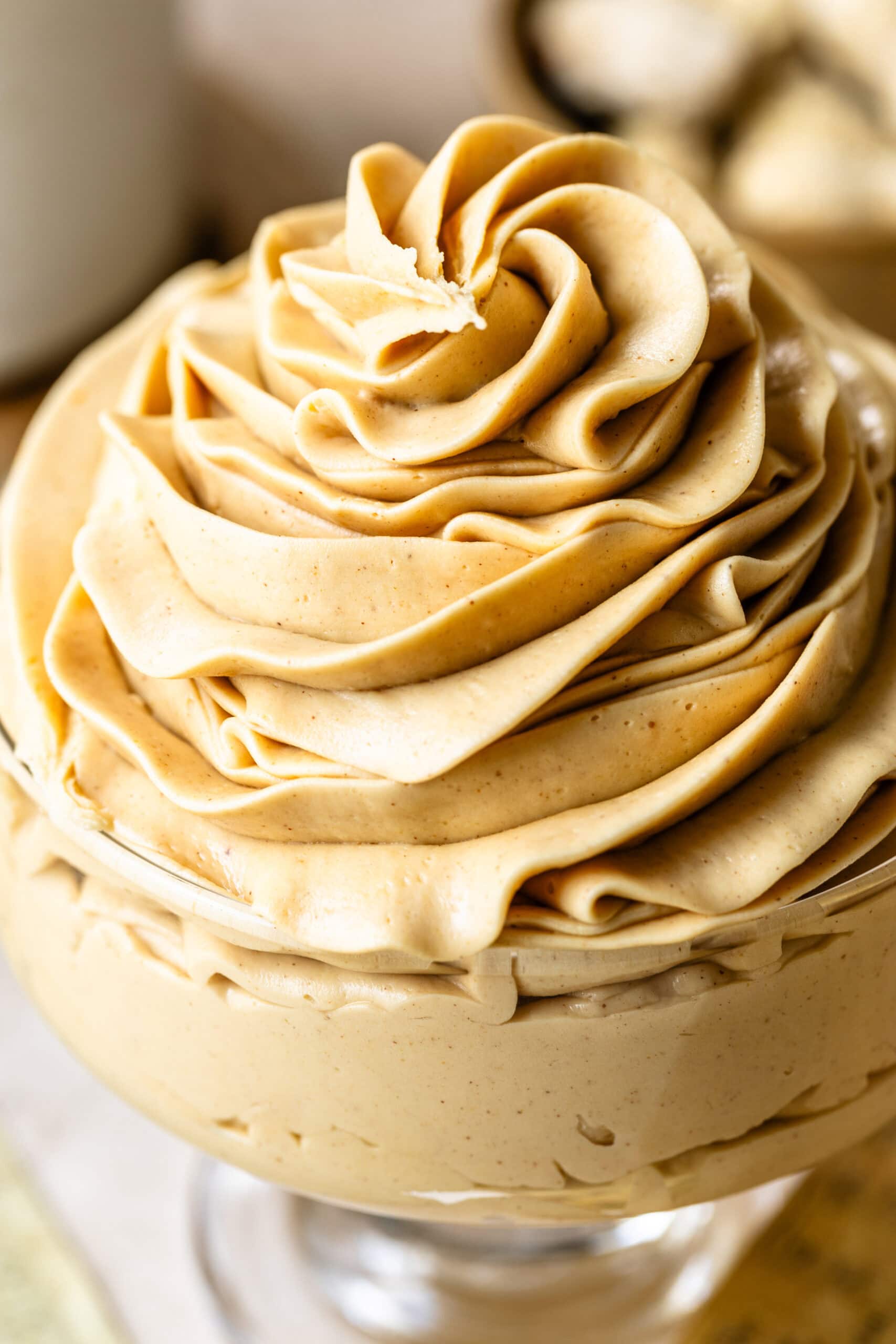 Peanut Butter Frosting Swirled in a Dish (Alternate Angle): Another close-up of the same glass dish filled with peanut butter frosting, taken from a slightly different angle. The frosting appears even more luscious, with its delicate swirls and rich, creamy consistency clearly visible. The soft focus on the background emphasizes the texture of the frosting.