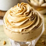 Peanut Butter Frosting in a Glass Dish: Close-up of a glass dessert dish filled with creamy, smooth peanut butter frosting. The frosting is beautifully swirled on top, showcasing its rich and fluffy texture. In the background, there are blurred peanuts and a white ceramic container, adding warmth and a homely feel to the scene.