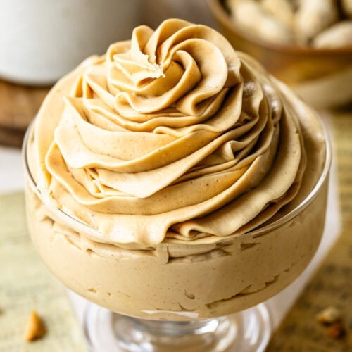 Peanut Butter Frosting in a Glass Dish: Close-up of a glass dessert dish filled with creamy, smooth peanut butter frosting. The frosting is beautifully swirled on top, showcasing its rich and fluffy texture. In the background, there are blurred peanuts and a white ceramic container, adding warmth and a homely feel to the scene.