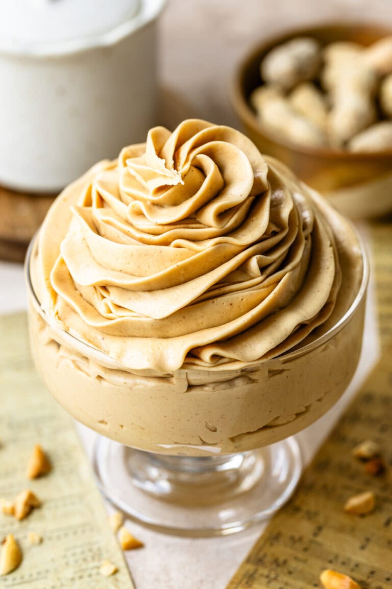 Peanut Butter Frosting in a Glass Dish: Close-up of a glass dessert dish filled with creamy, smooth peanut butter frosting. The frosting is beautifully swirled on top, showcasing its rich and fluffy texture. In the background, there are blurred peanuts and a white ceramic container, adding warmth and a homely feel to the scene.