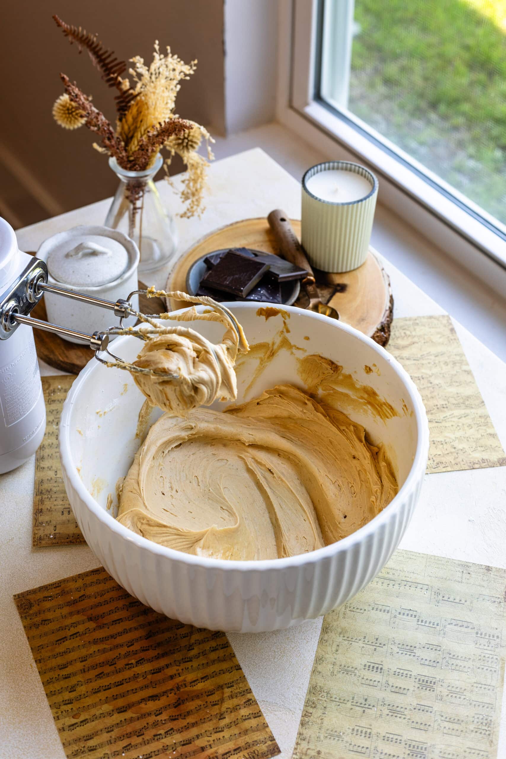 Mixing Peanut Butter Frosting in a Bowl: A large white mixing bowl filled with freshly made peanut butter frosting, with a hand mixer still resting on top. The beaters are coated in the creamy frosting, and the bowl sits on a table near a bright window. The background features a vase with dried flowers, a lit candle, and some pieces of chocolate on a wooden tray, creating a cozy kitchen atmosphere.