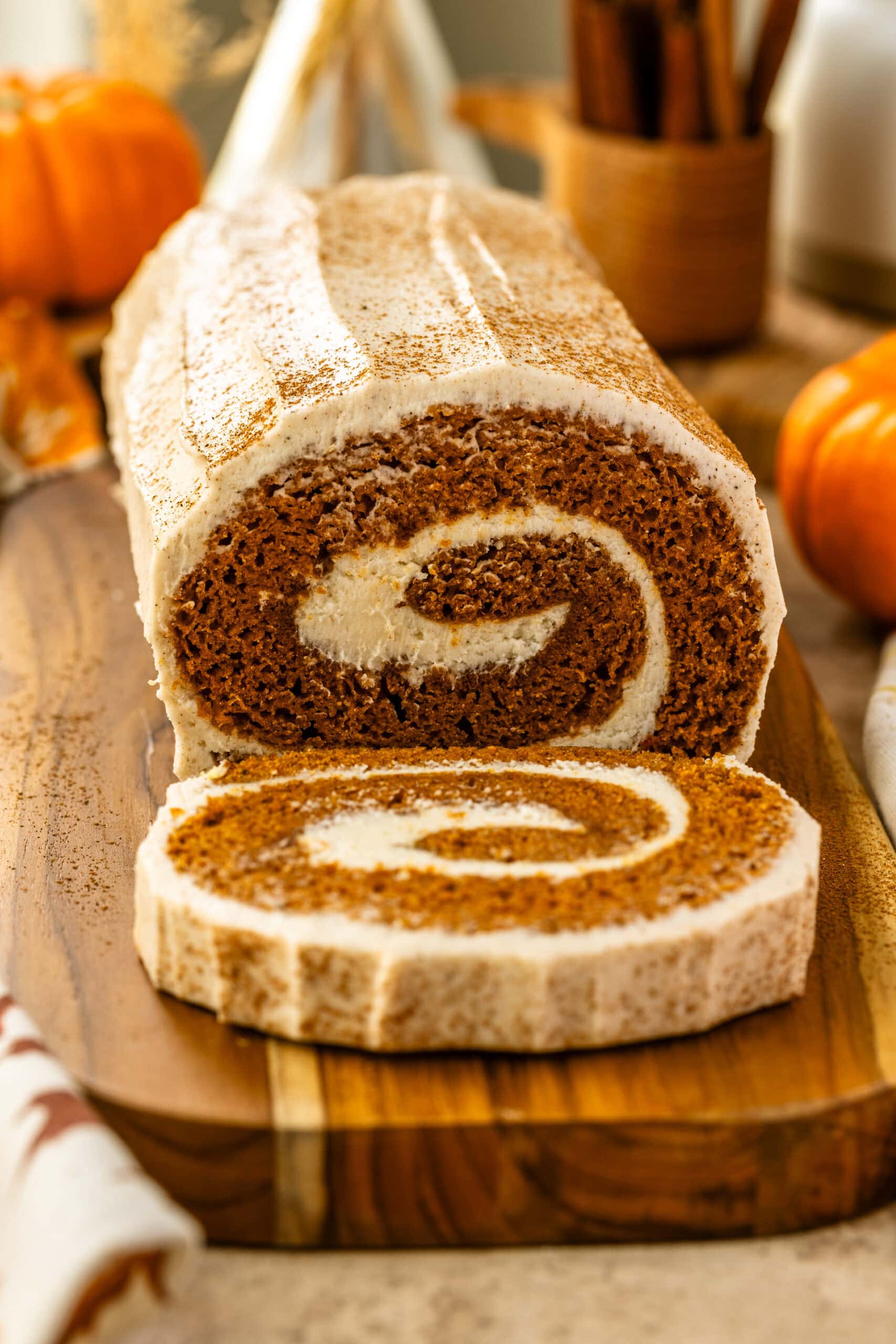 A close-up view of the frosted pumpkin roll cake on a wooden board. The swirl of the cream cheese filling is visible inside the cake, with the cinnamon-topped frosting on the surface. Autumn decorations, such as pumpkins and dried flowers, surround the scene.