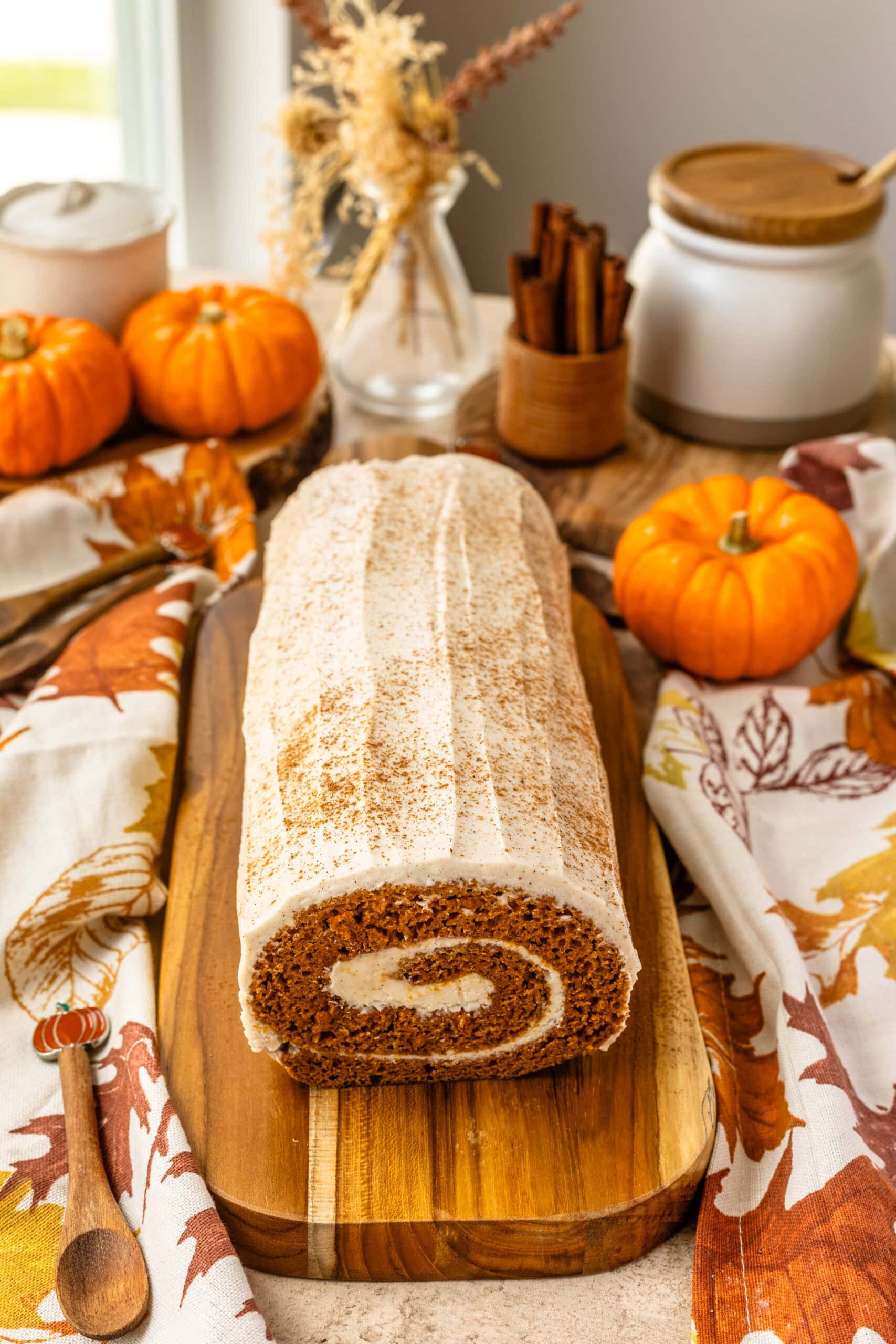 A frontal view of the entire pumpkin roll cake, highlighting the swirl of the cream cheese filling and the smooth frosting on top. The cake is dusted with cinnamon, and the background is decorated with fall-themed elements, including pumpkins, a vase of dried flowers, and cinnamon sticks.