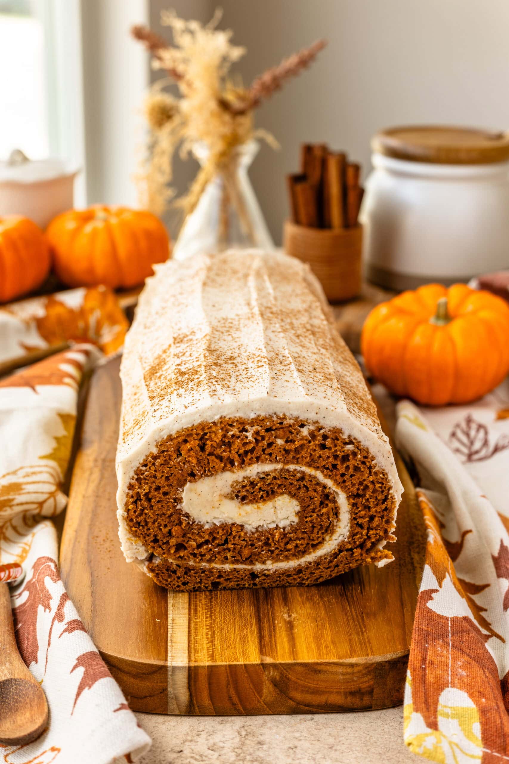 A frontal view of the entire pumpkin roll cake, highlighting the swirl of the cream cheese filling and the smooth frosting on top. The cake is dusted with cinnamon, and the background is decorated with fall-themed elements, including pumpkins, a vase of dried flowers, and cinnamon sticks.
