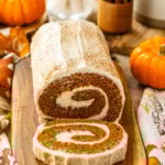 A close-up view of the frosted pumpkin roll cake on a wooden board. The swirl of the cream cheese filling is visible inside the cake, with the cinnamon-topped frosting on the surface. Autumn decorations, such as pumpkins and dried flowers, surround the scene.