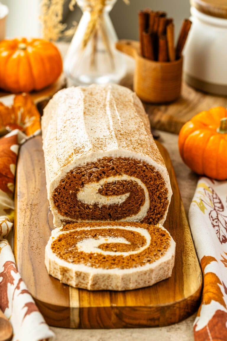 A close-up view of the frosted pumpkin roll cake on a wooden board. The swirl of the cream cheese filling is visible inside the cake, with the cinnamon-topped frosting on the surface. Autumn decorations, such as pumpkins and dried flowers, surround the scene.