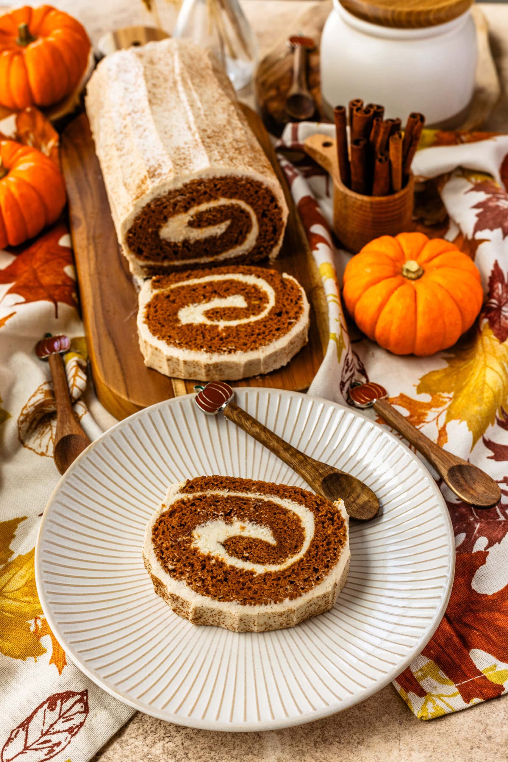 A sliced pumpkin roll cake with cream cheese frosting is placed on a wooden board. Two slices are served on a decorative white plate, showcasing the spiral of the filling. Small pumpkins and cinnamon sticks are seen as part of the autumn-themed decor.