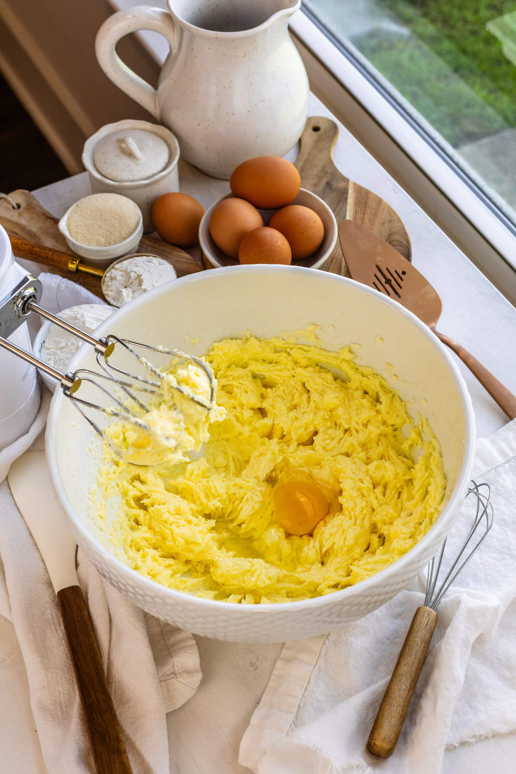 Egg being added to a butter and sugar mixture in a mixing bowl, with eggs, sugar, flour, and kitchen tools on the counter in the background.
