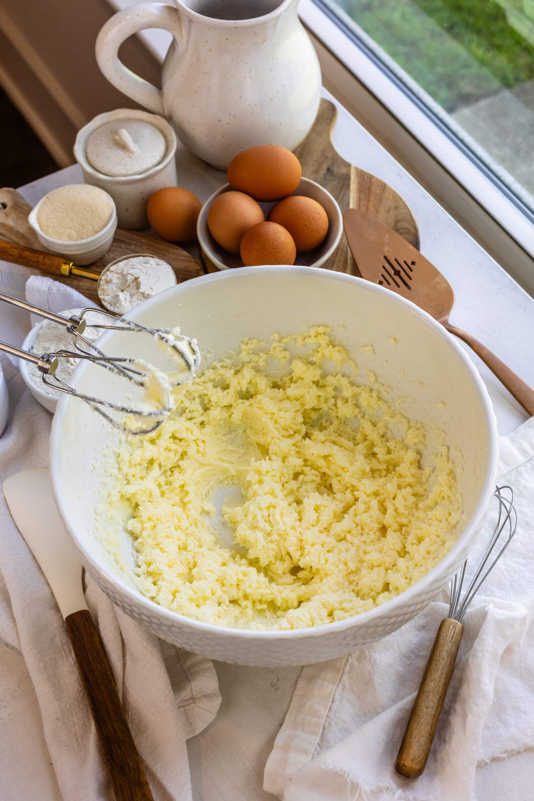 Butter and sugar being creamed together in a mixing bowl with eggs, flour, and other baking ingredients set around the kitchen counter.