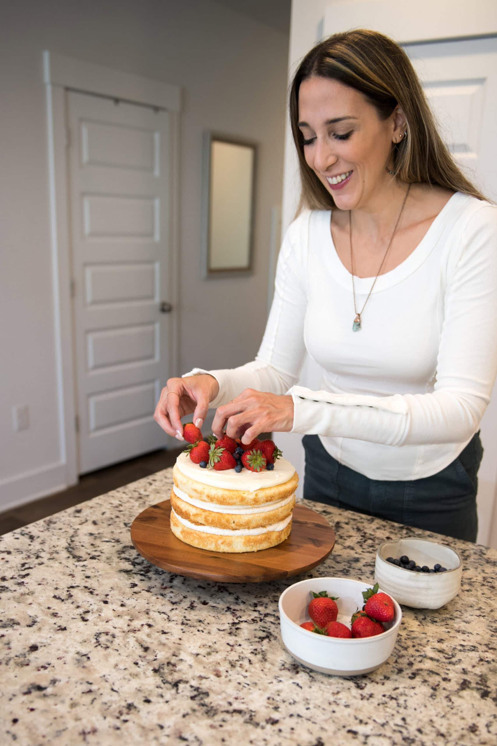 A woman decorating a naked vanilla layer cake with fresh strawberries and blueberries on a kitchen counter, with bowls of berries nearby.