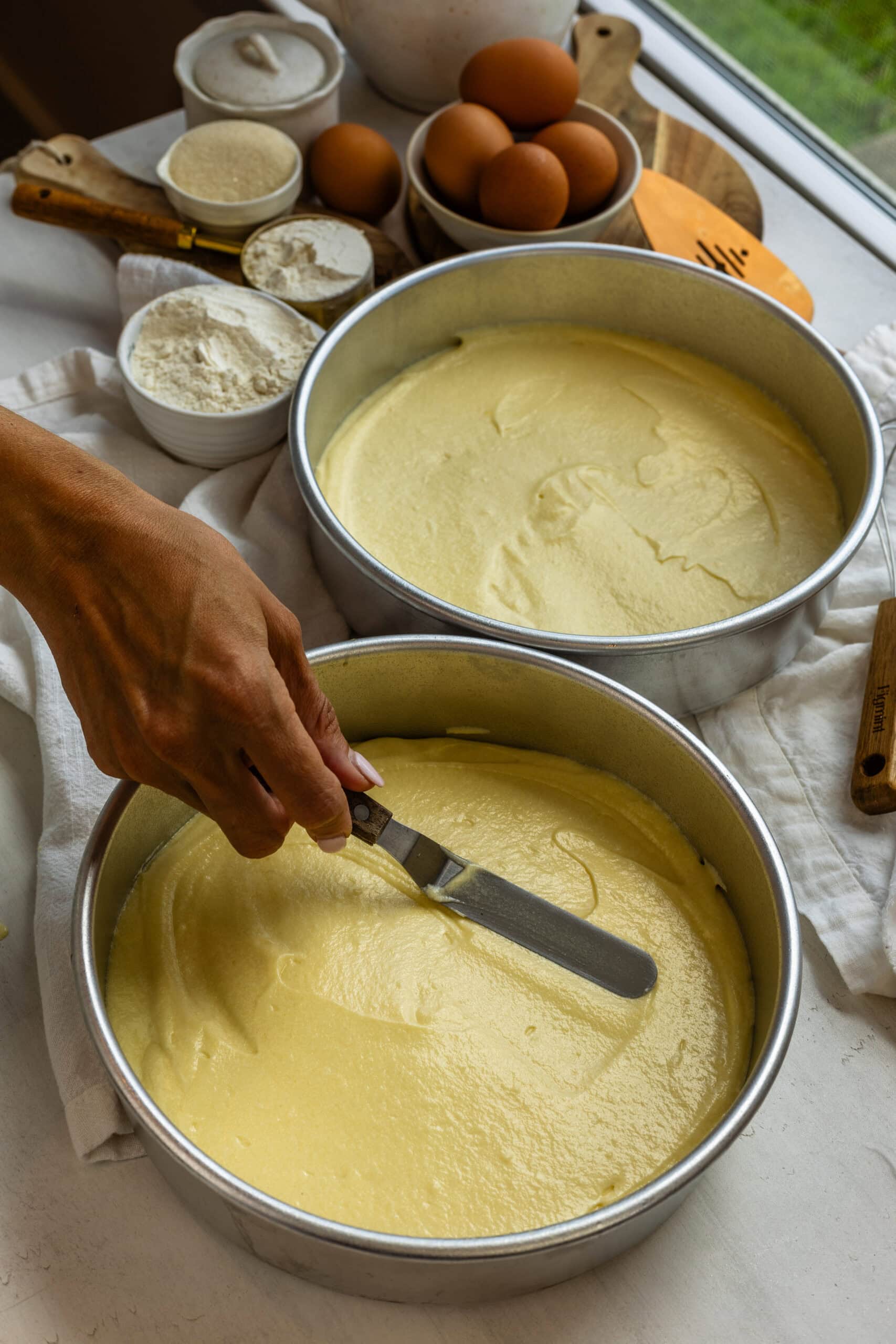 Batter being spread evenly in round cake pans with an offset spatula, with flour, eggs, and other baking ingredients visible on the counter in the background.