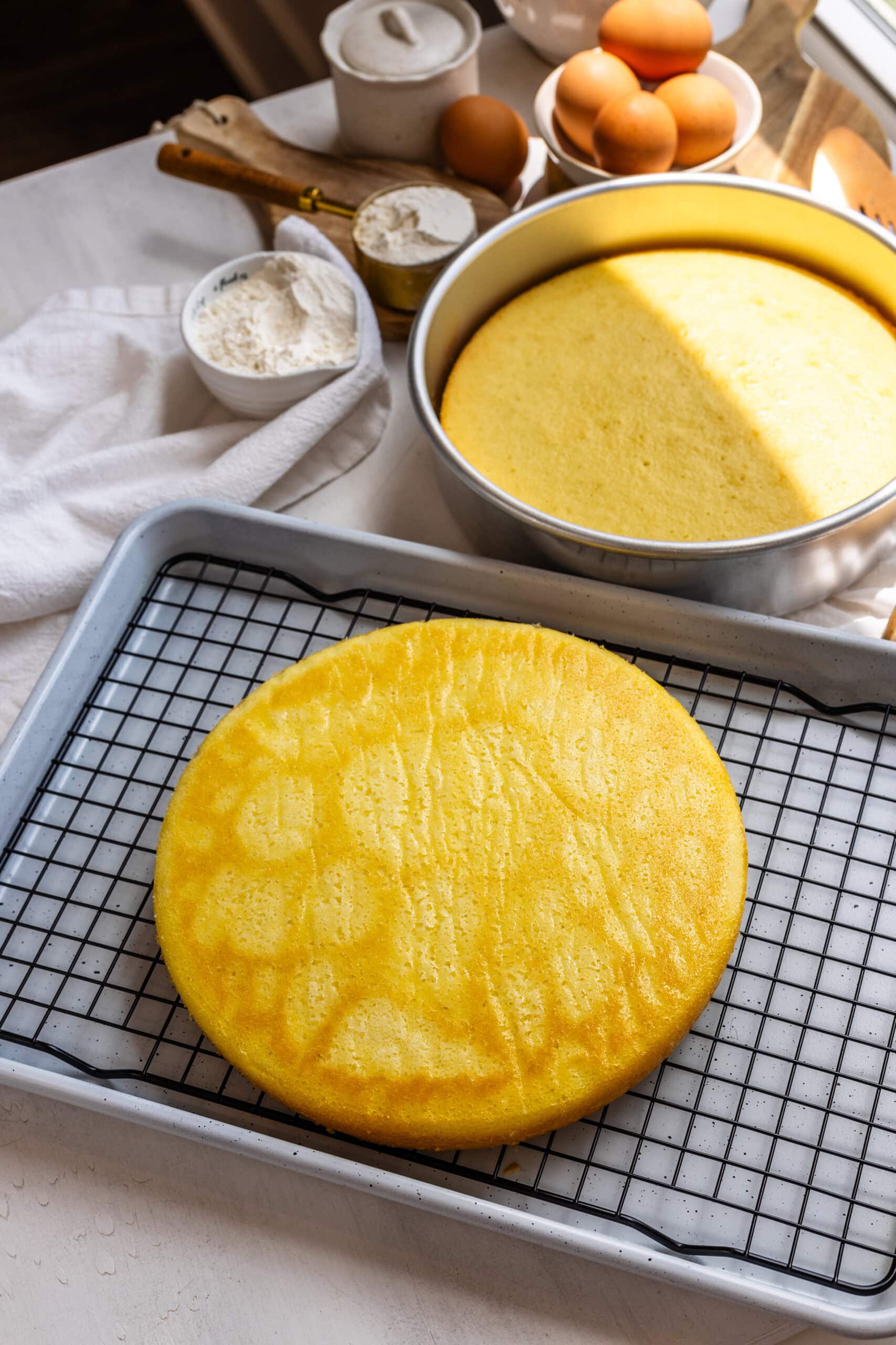 Freshly baked vanilla cake layers cooling on a wire rack, with baking ingredients like flour, eggs, and sugar in the background.