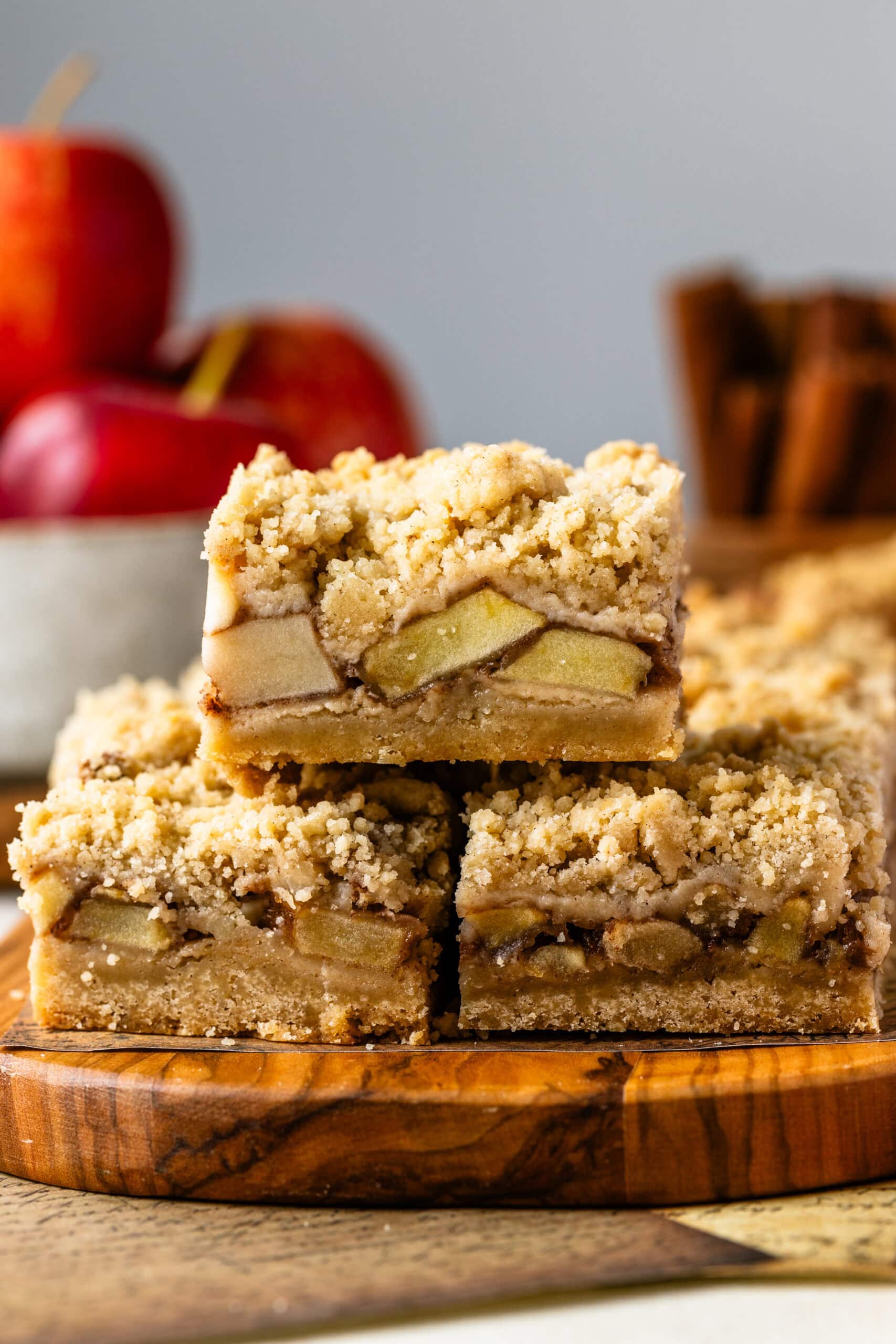 The image features a stack of golden apple crumble bars. The bars have a crunchy, crumbly top and reveal tender spiced apples nestled between the layers. The third image presents the bars neatly cut and arranged on a parchment-lined wooden board, highlighting their even, golden-brown crust. Surrounding the bars are apples, cinnamon sticks, and brown sugar, emphasizing the warm, autumnal flavors of the dessert.