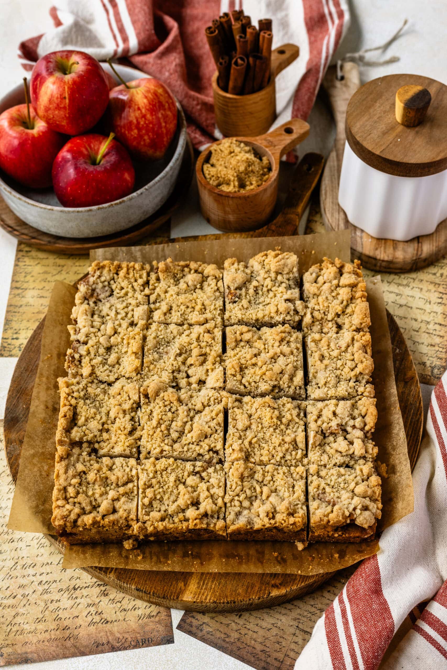 The image features freshly baked apple crumb bars neatly cut into squares, placed on a wooden board lined with parchment paper. The bars have a golden, crumbly topping, showcasing their inviting texture. Surrounding the bars are elements that highlight the ingredients used, including a bowl of red apples, a jar of cinnamon sticks, and a wooden container of brown sugar, creating a warm, cozy autumnal vibe. The scene is set against a rustic background, with handwritten notes and a striped kitchen towel, enhancing the homemade and comforting feel of the dessert.