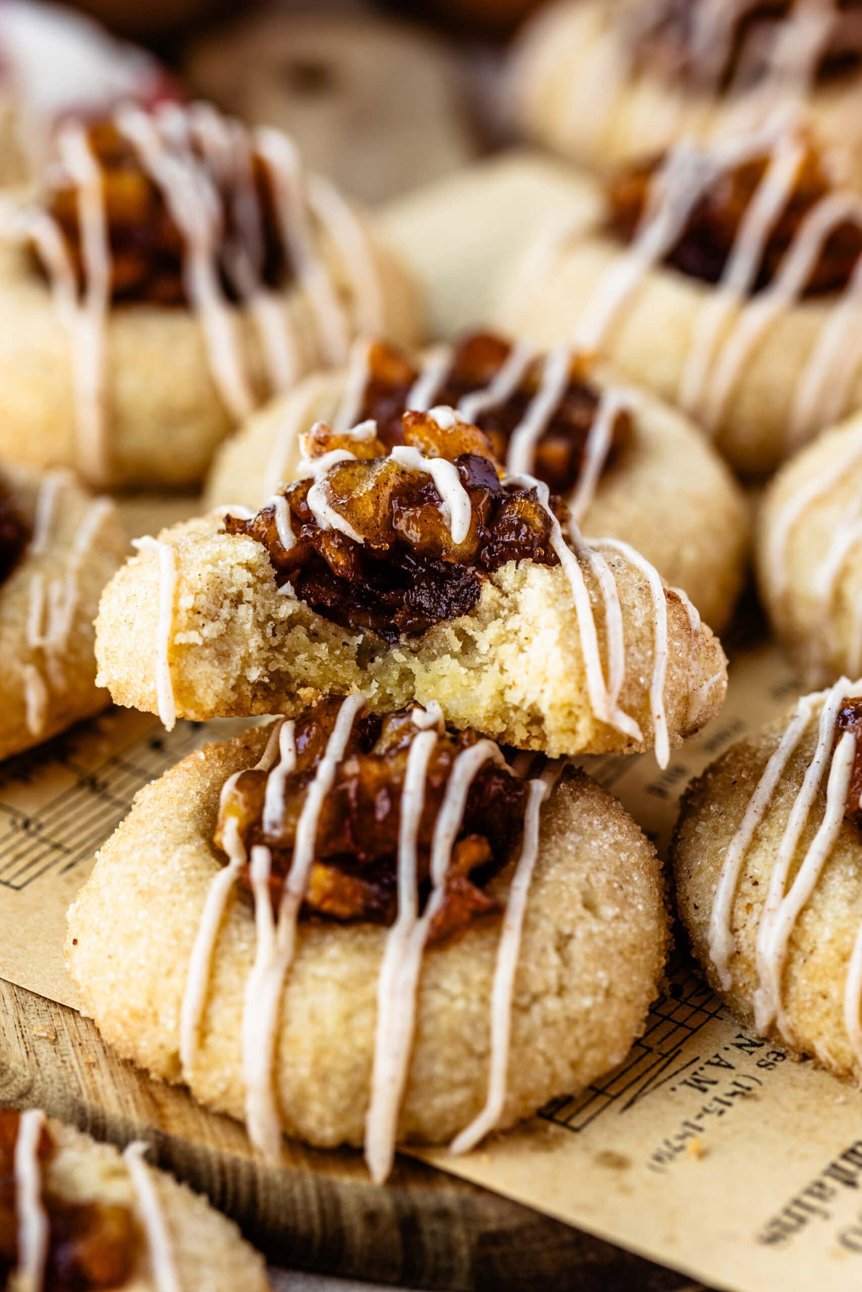 A close-up of apple pie cookies stacked on a rustic board, with one cookie featuring a bite taken out, revealing its soft, tender interior. Each cookie is topped with a rich, caramelized apple filling and drizzled with a cinnamon glaze, highlighting their texture and autumnal flavors. The combination of the golden cookie, glossy apple topping, and delicate glaze drizzle creates an inviting, warm look, perfect for fall festivities.