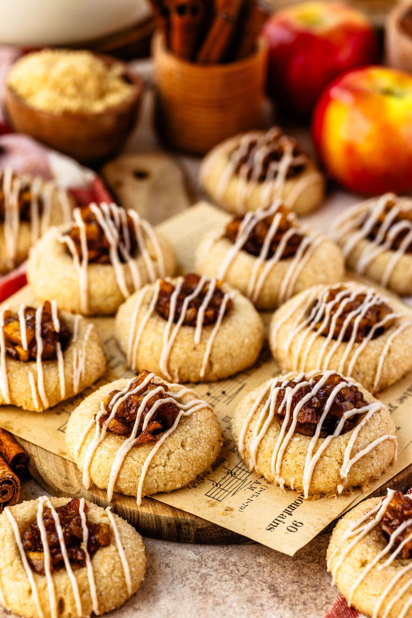 A close-up of freshly baked apple pie cookies arranged on a wooden board, each cookie topped with caramelized apple filling and drizzled with a cinnamon-spiced glaze. The glaze adds a decorative, festive touch to the cookies, complementing their golden-brown color and sugared coating. Fresh apples, cinnamon sticks, and brown sugar in the background enhance the warm, cozy, autumnal setting.