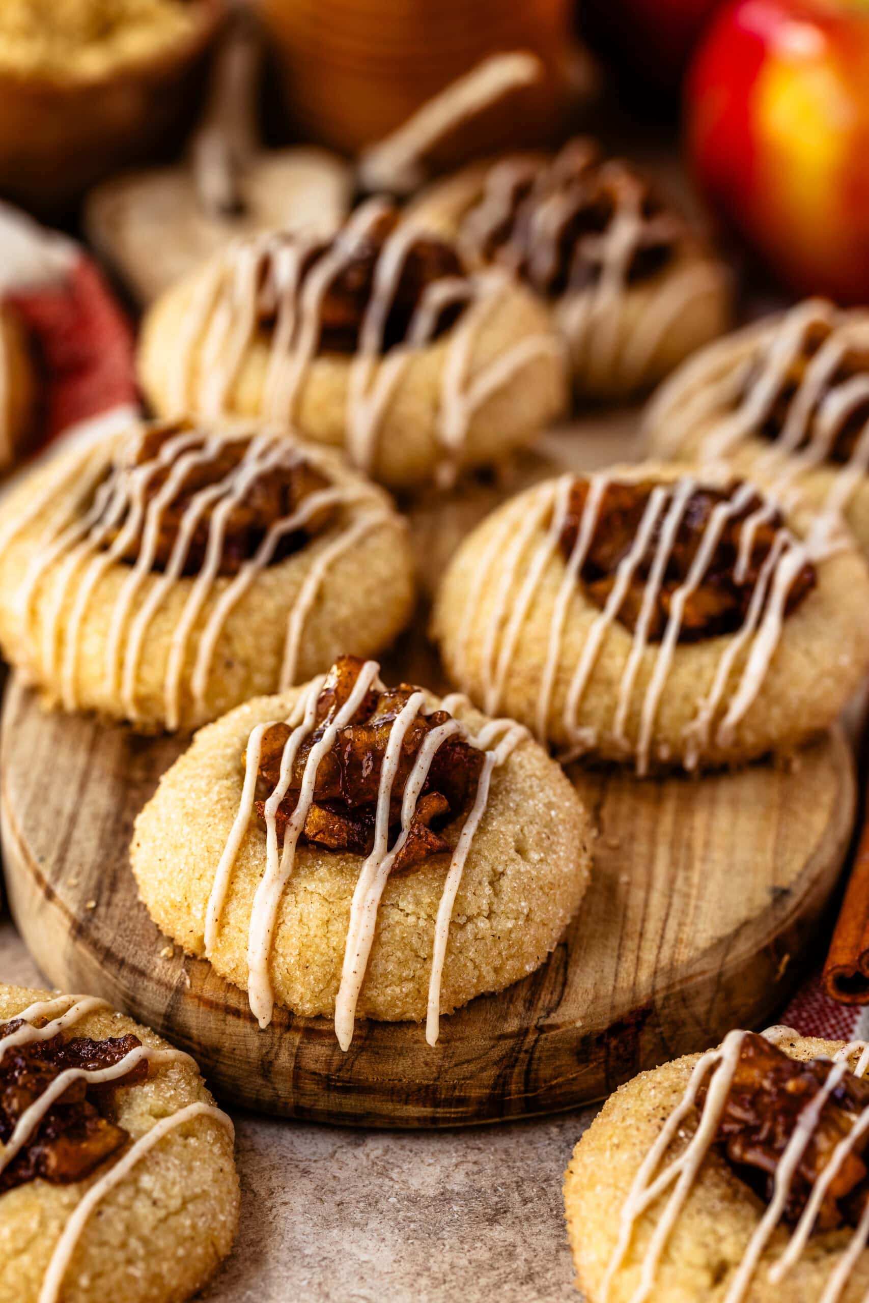 A close-up of freshly baked apple pie cookies arranged on a wooden board, each cookie topped with caramelized apple filling and drizzled with a cinnamon-spiced glaze. The glaze adds a decorative, festive touch to the cookies, complementing their golden-brown color and sugared coating. Fresh apples, cinnamon sticks, and brown sugar in the background enhance the warm, cozy, autumnal setting.