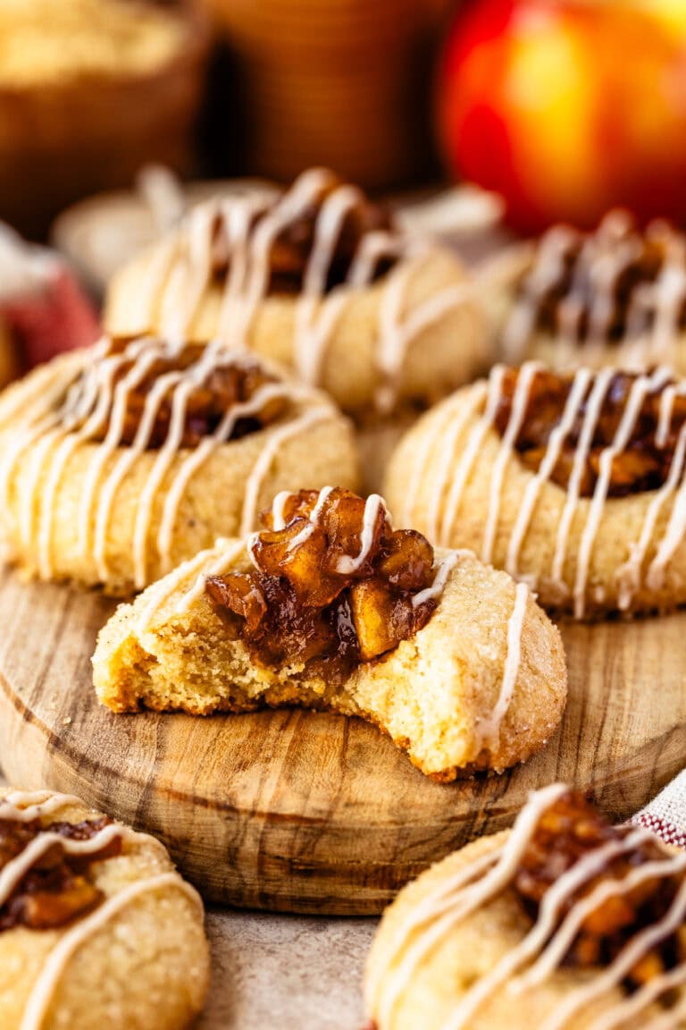 A close-up of apple pie cookies with a focus on one cookie that has a bite taken out, revealing a soft, tender interior. Each cookie is topped with a rich caramelized apple filling and drizzled with a cinnamon-spiced glaze, enhancing its warm, sweet flavor. The cookies are displayed on a wooden board, surrounded by others, capturing their inviting, homemade texture. The vibrant apple filling and glaze drizzle add an autumnal charm, making these cookies look as comforting as they are delicious.